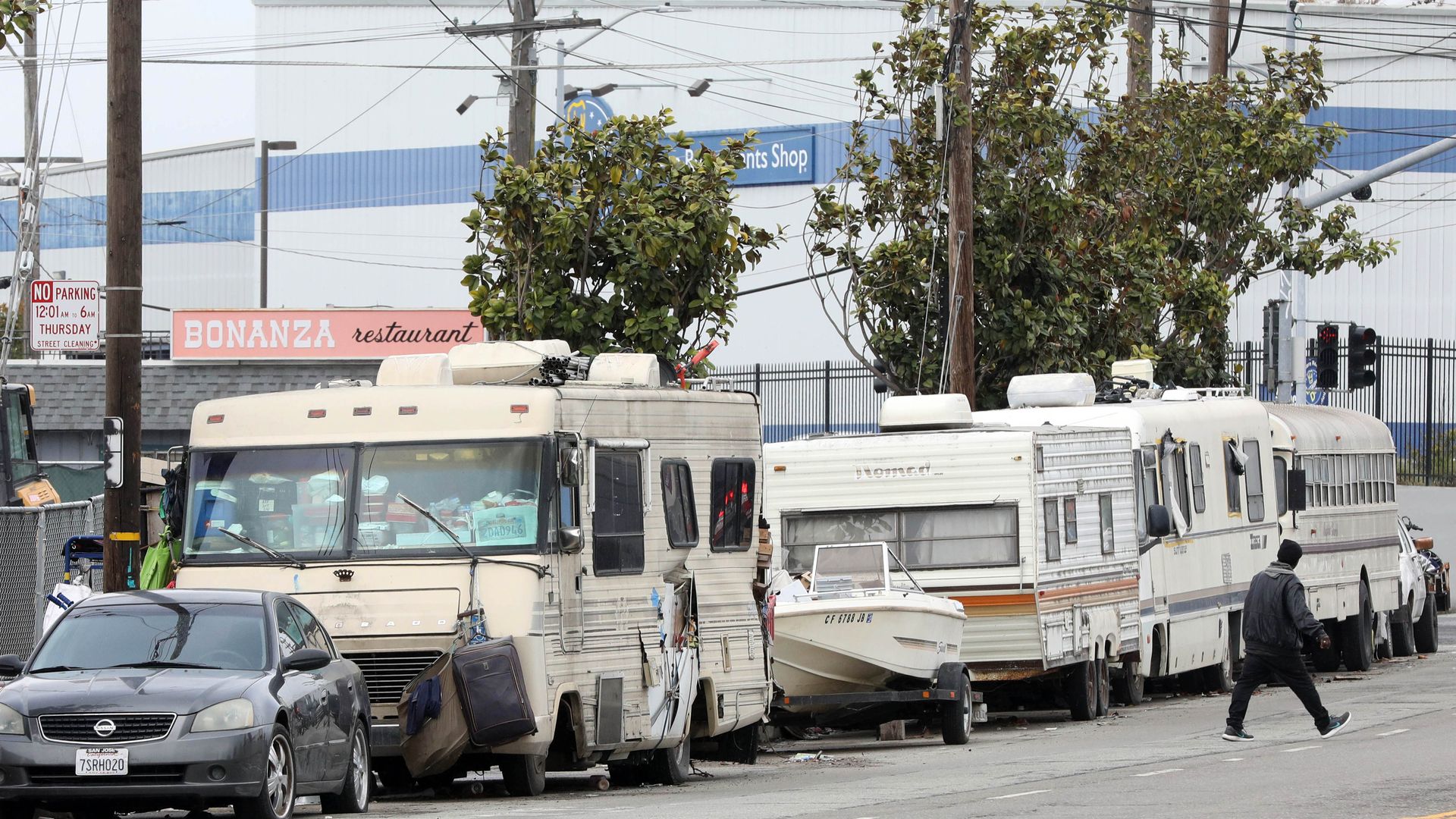 Line of beige and white RVs and a small boat parked on a city street near trees and buildings, with a black car and a person crossing the road in a gray jacket and hat.