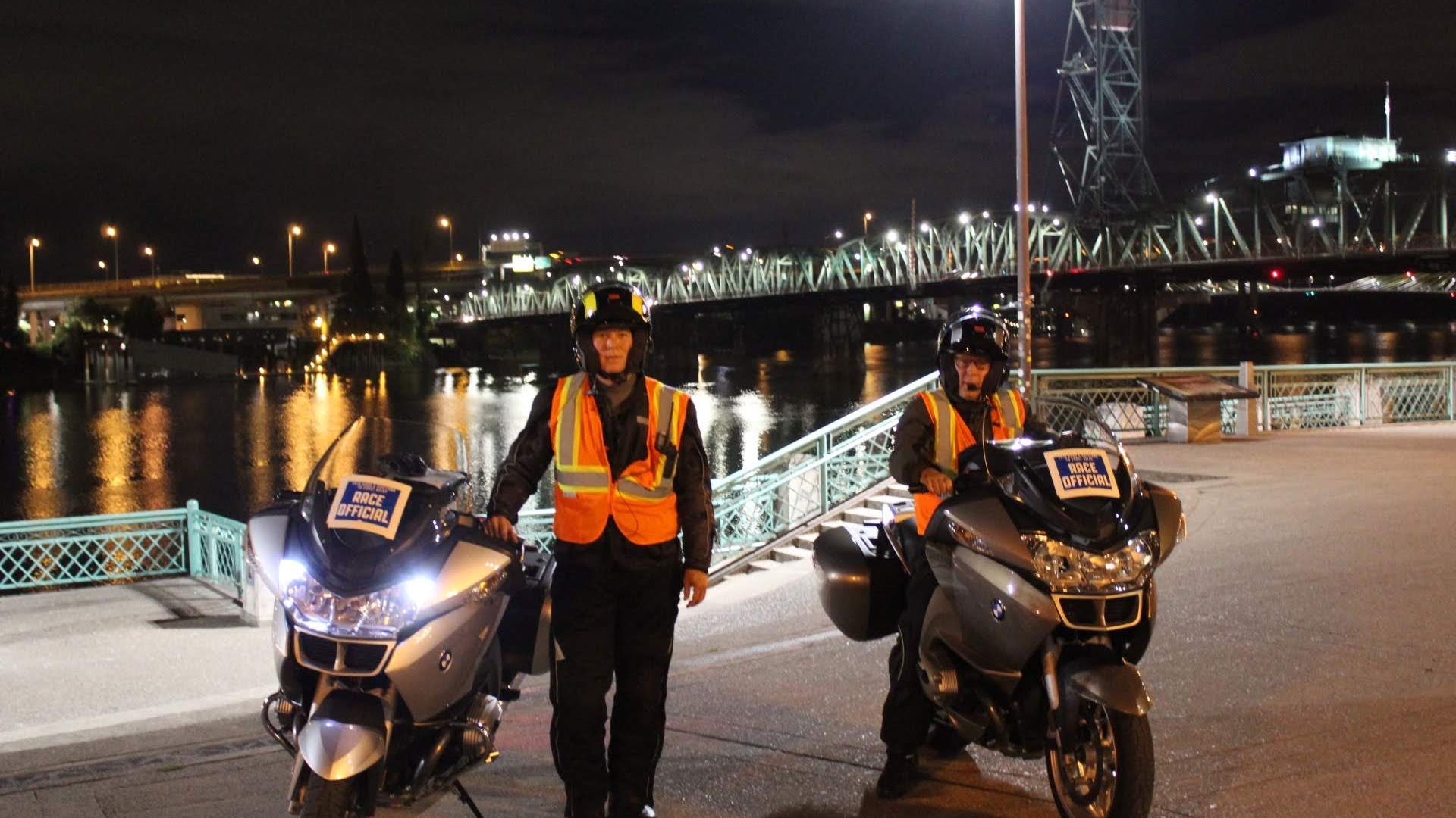 Night scene on a riverside with two motorcycle officers in orange reflective vests standing near their bikes; a tall steel bridge lit by floodlights spans the water, reflections shimmer on the river.