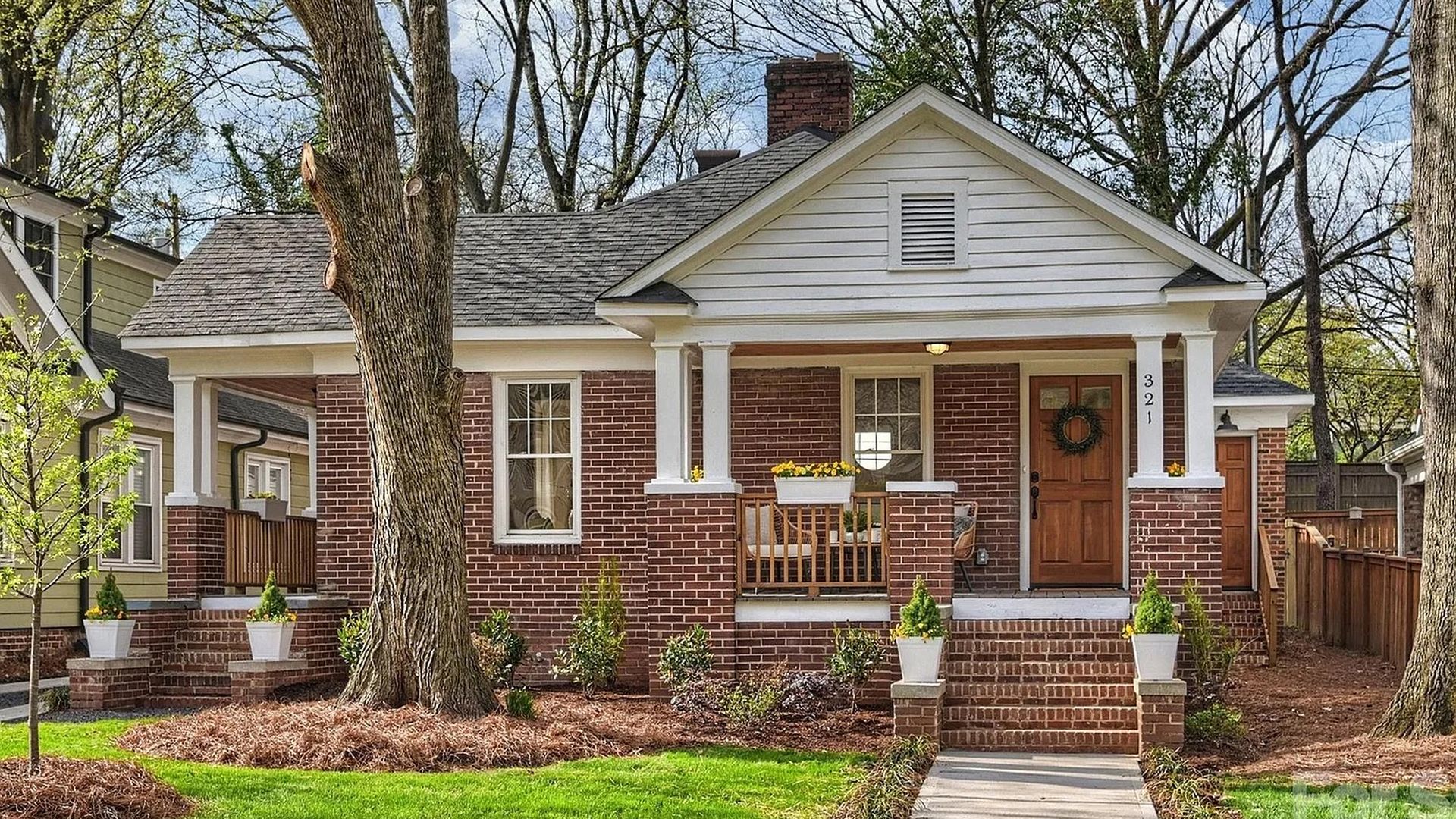 Brick cottage with white trim and a front porch supported by two columns. Wooden door with a wreath, brick steps, and planters with yellow flowers; green lawn, large trees, and blue sky.