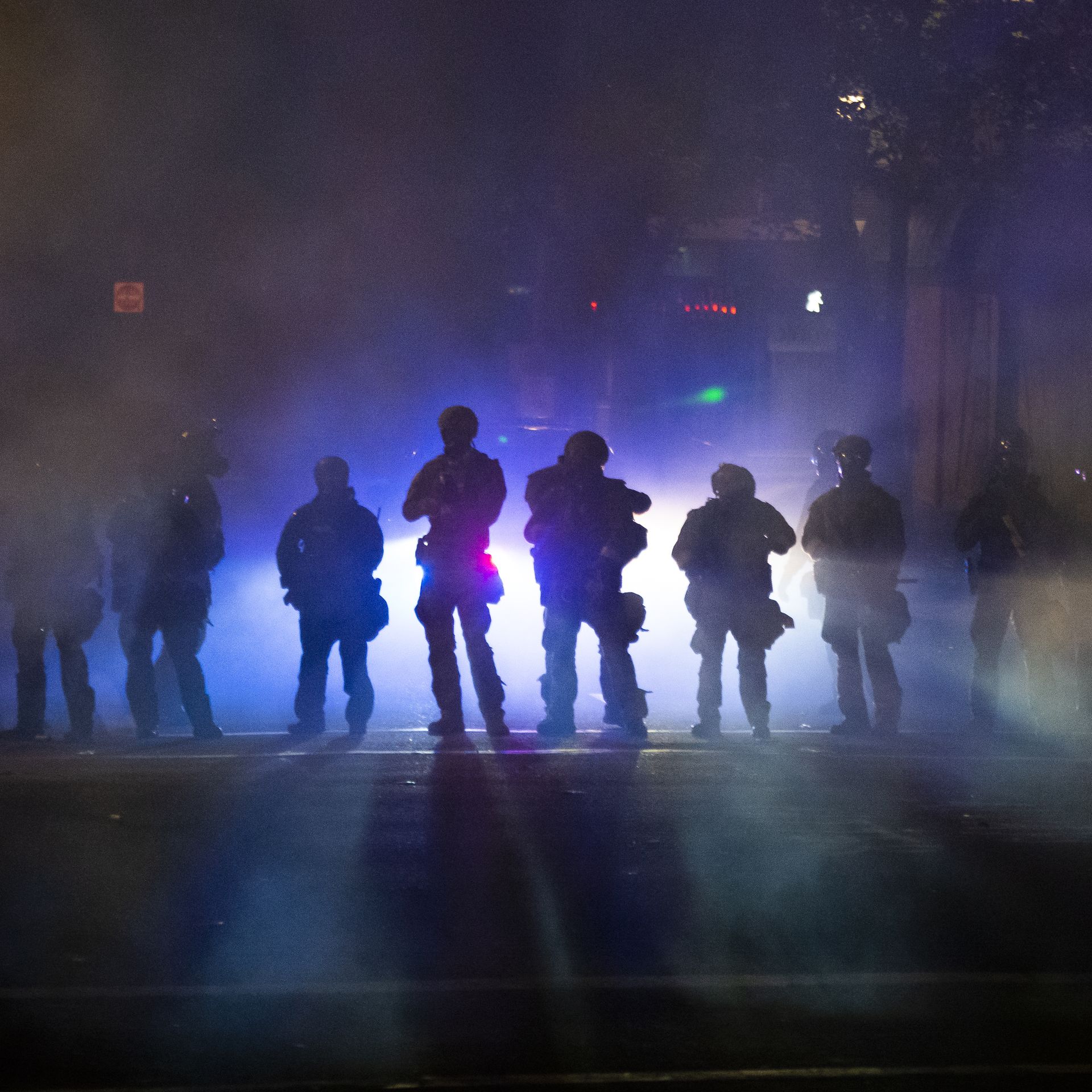A line of armed officers are backlit by police lights and tear gas