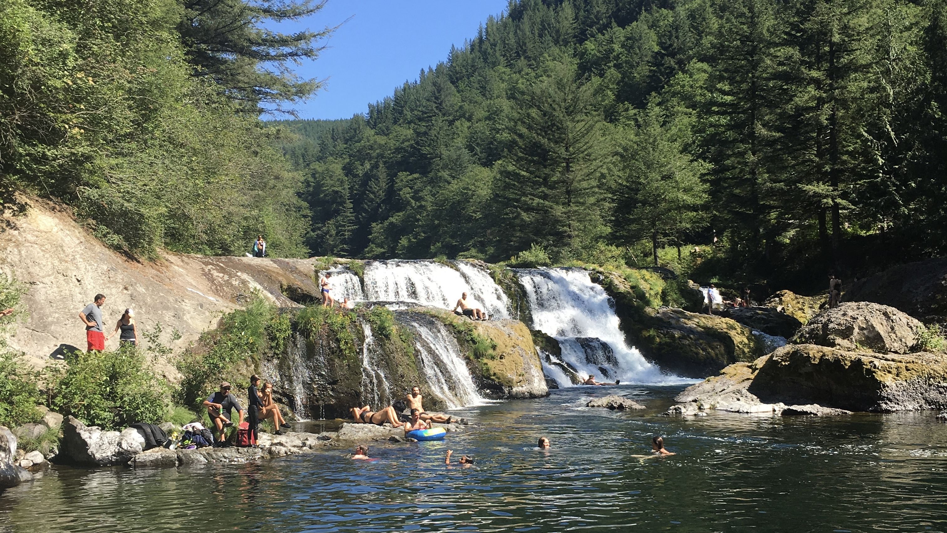 People enjoying a sunny day swimming and relaxing by a wide waterfall surrounded by rocky banks and dense green forest under a clear blue sky.