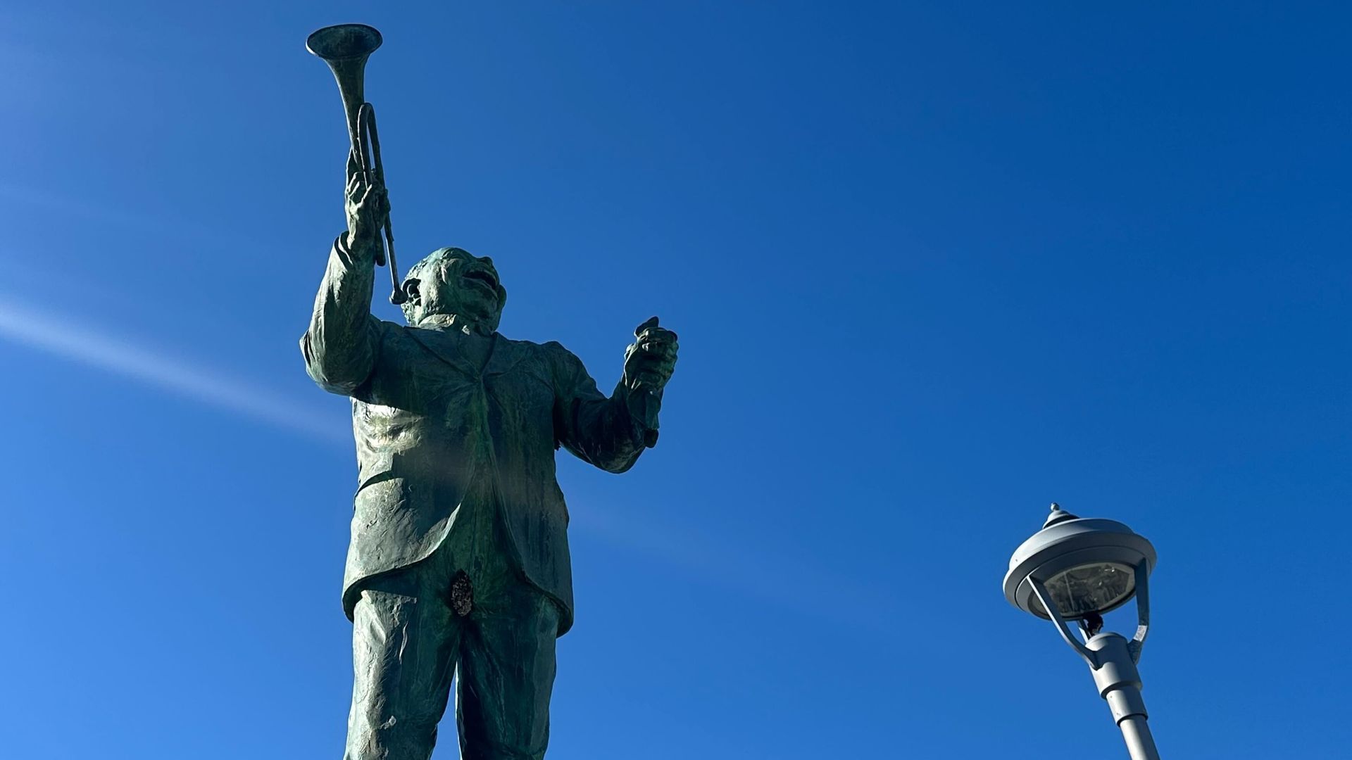 Bronze statue of a man in a suit holding a trumpet in one hand and a small object in the other, set against a clear blue sky beside a tall streetlamp.