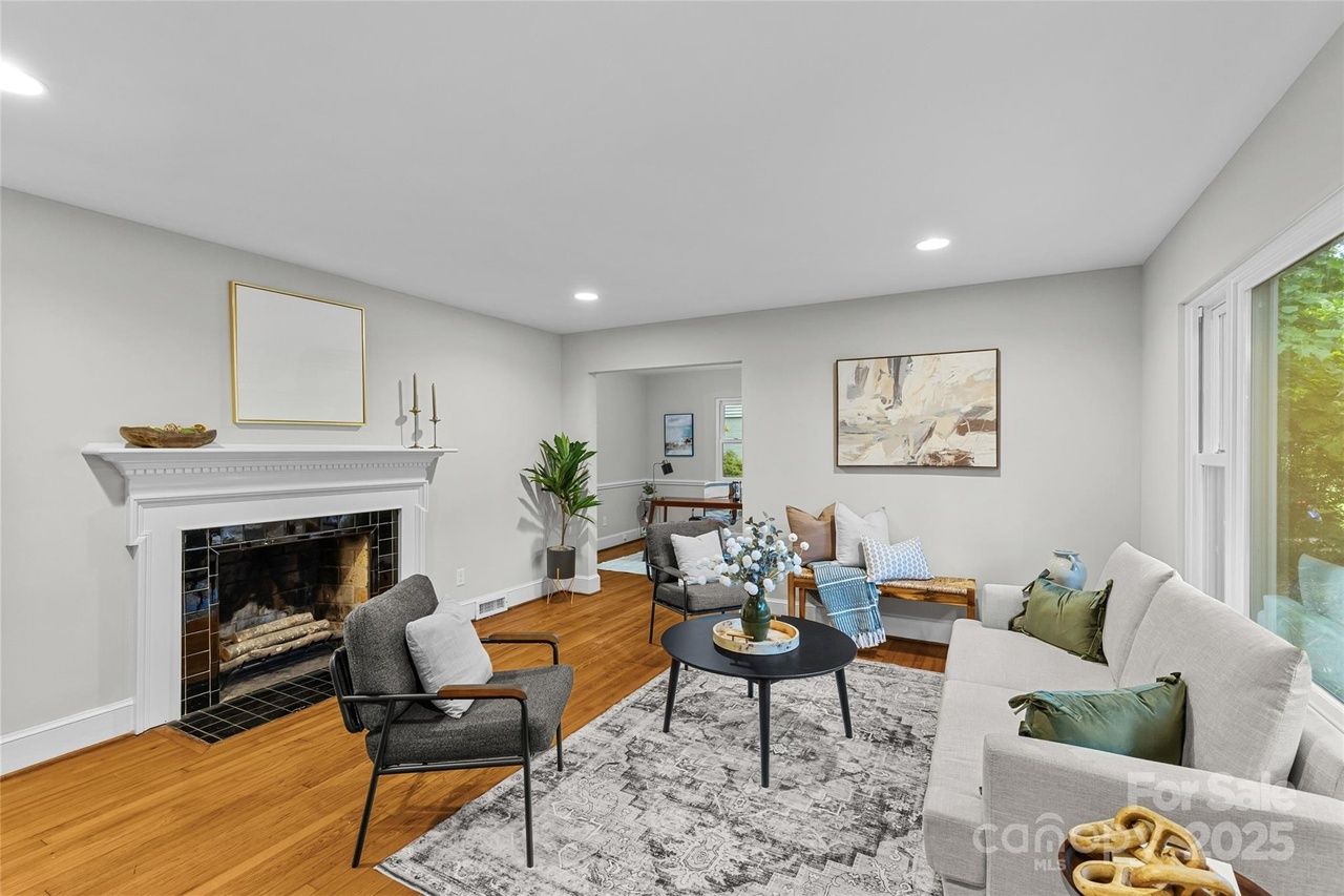 Bright living room with light gray walls, wood floor, white fireplace, gray armchairs, beige sofa with green pillows, black round coffee table, patterned rug, and large window showing greenery.