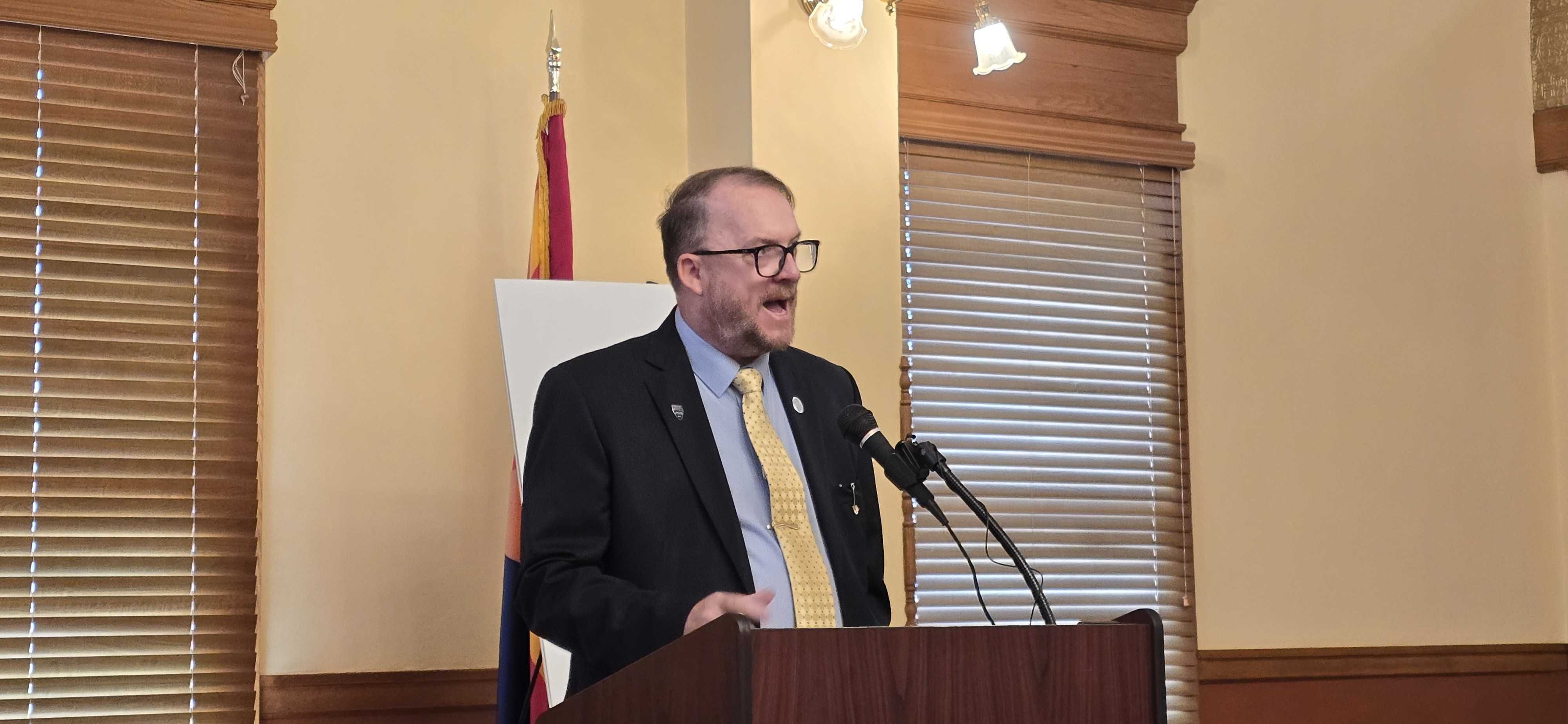 Frederick Tack in a dark suit, light blue shirt, yellow tie, and glasses speaks at a podium with a microphone, in a room with wooden blinds and a flag behind him.
