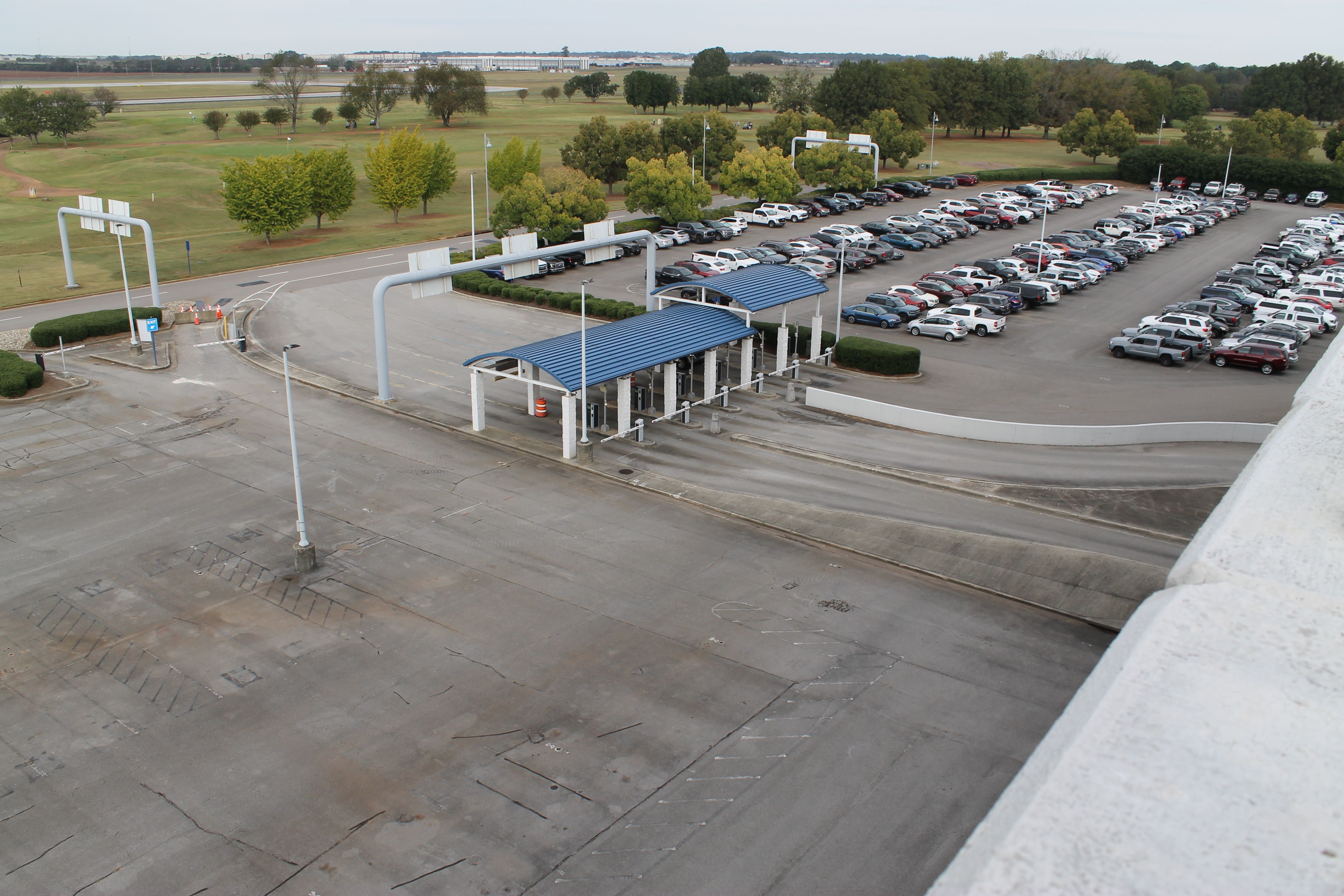A large parking lot with many cars parked in rows next to a blue-roofed toll or checkpoint area, surrounded by green trees and open grassy fields under a cloudy sky.