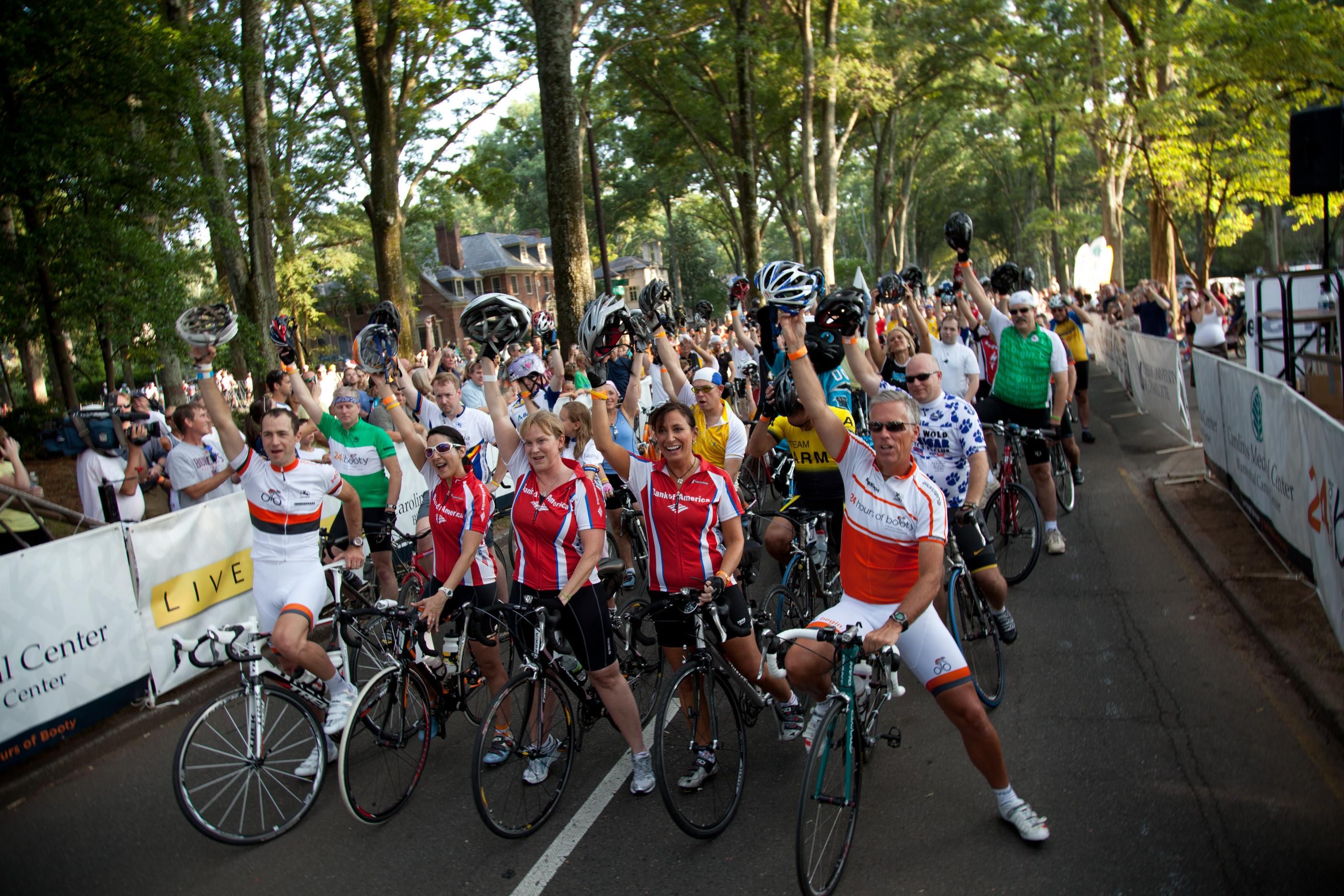A large group of cyclists gathered on a tree-lined street hold their helmets up, wearing colorful cycling jerseys, ready for an event or race with spectators nearby.
