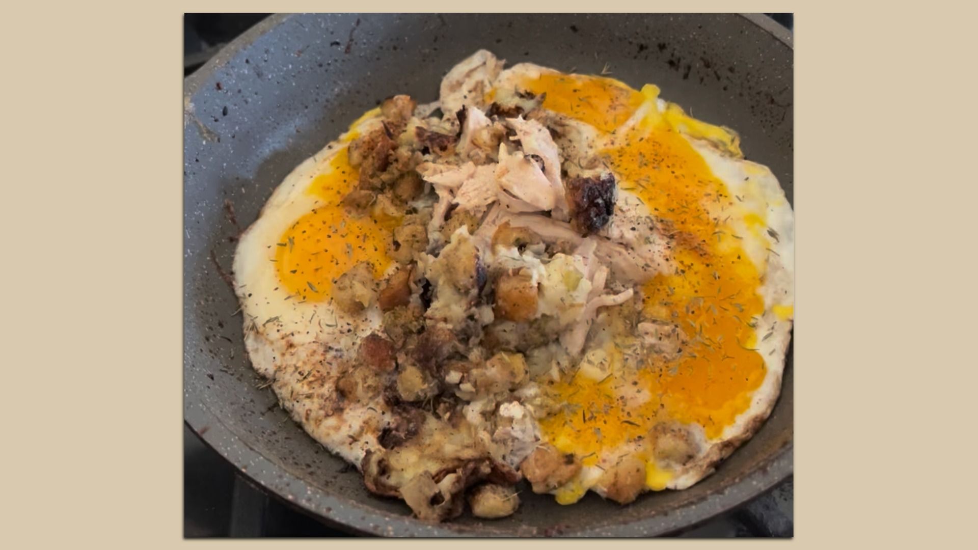 Two sunny-side-up eggs cooking in a gray speckled pan topped with seasoned cooked turkey and breadcrumb stuffing, sprinkled with herbs.