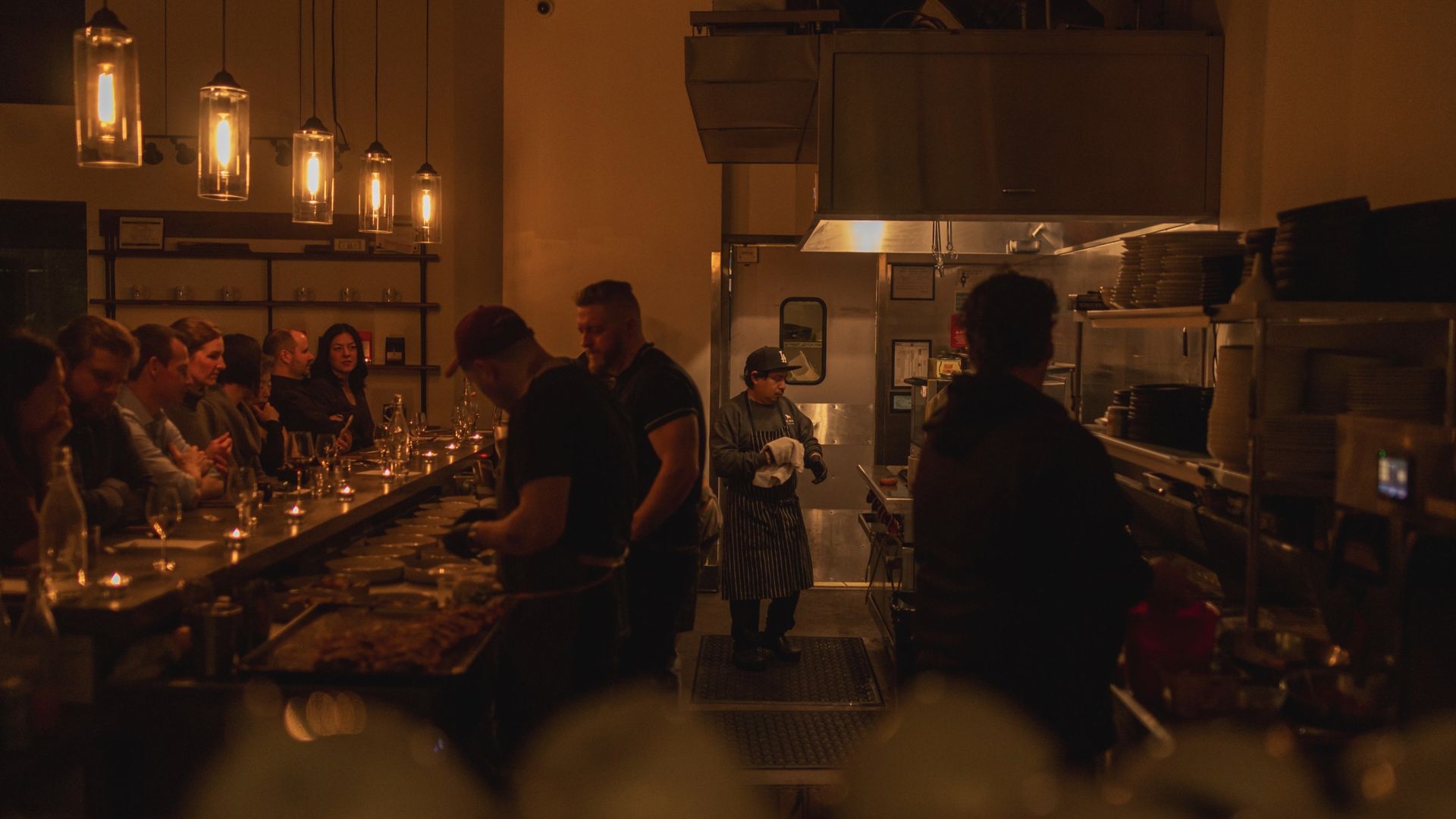 Dimly lit restaurant kitchen and dining counter with chefs preparing food and customers seated, warm overhead pendant lights, plates, glasses, and utensils visible.