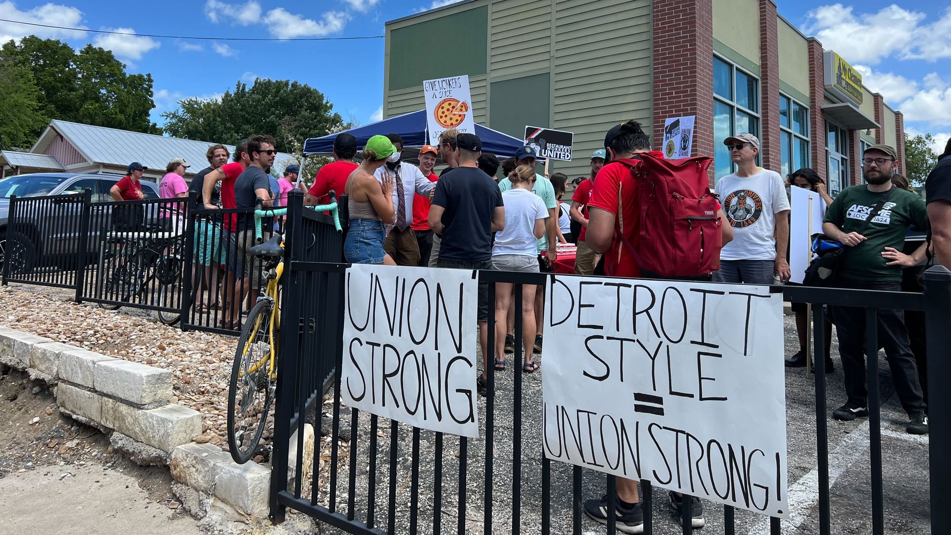 People gather outside near a black fence with two posters that read "union strong" and "Detroit style=union strong"