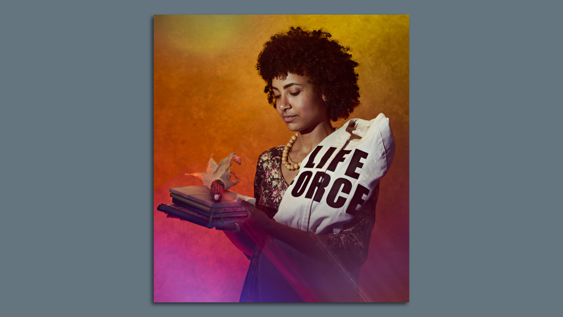 A woman holding corn on some books and a white cloth that reads LIFE FORCE on her shoulder. 