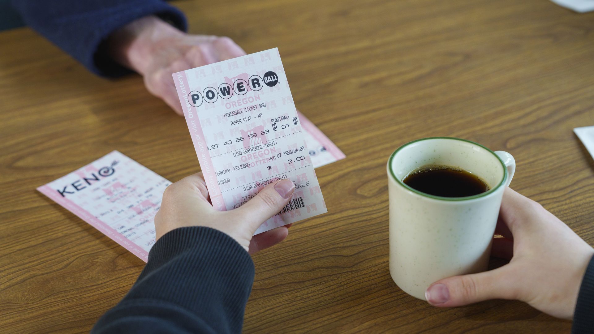 Han holding lottery tickets and coffee cup at a diner table with another hand pushing a ticket across the table
