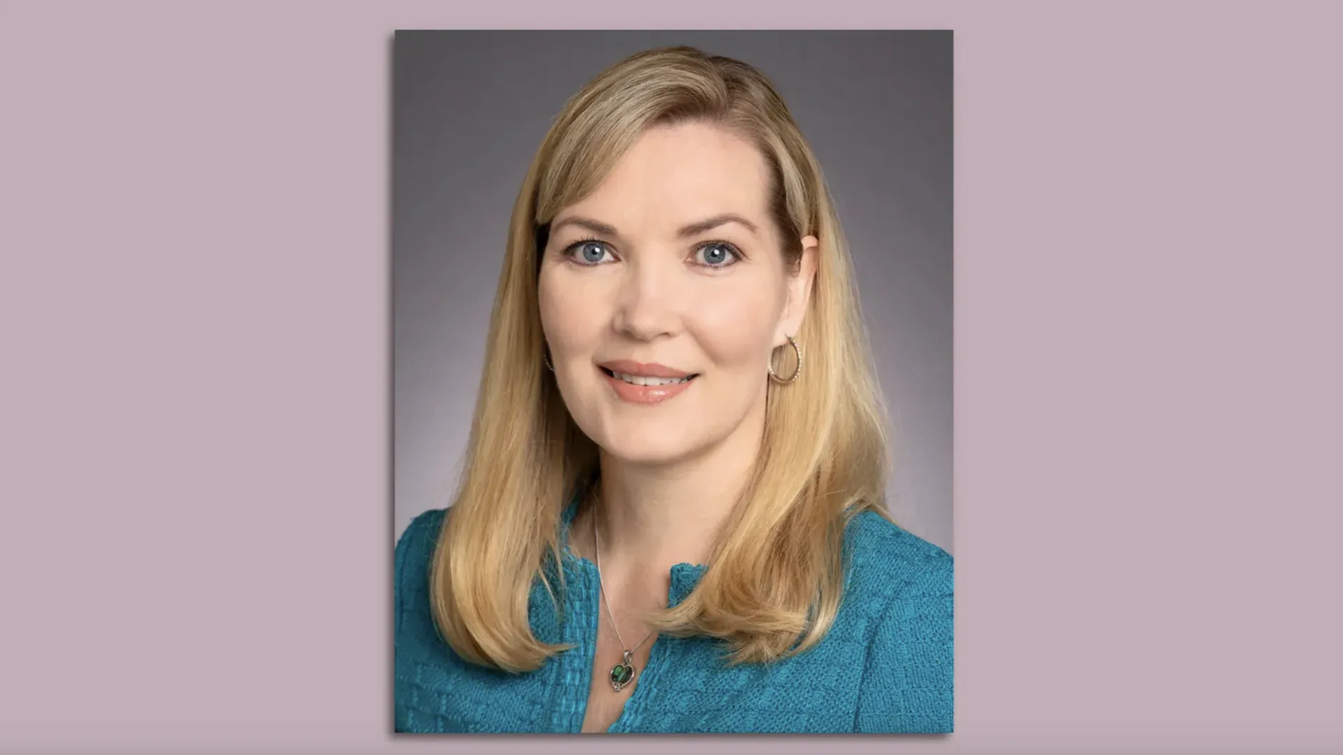 Portrait of a smiling blonde woman with blue eyes, wearing a teal blouse, hoop earrings, and a heart pendant necklace against a gray gradient backdrop.