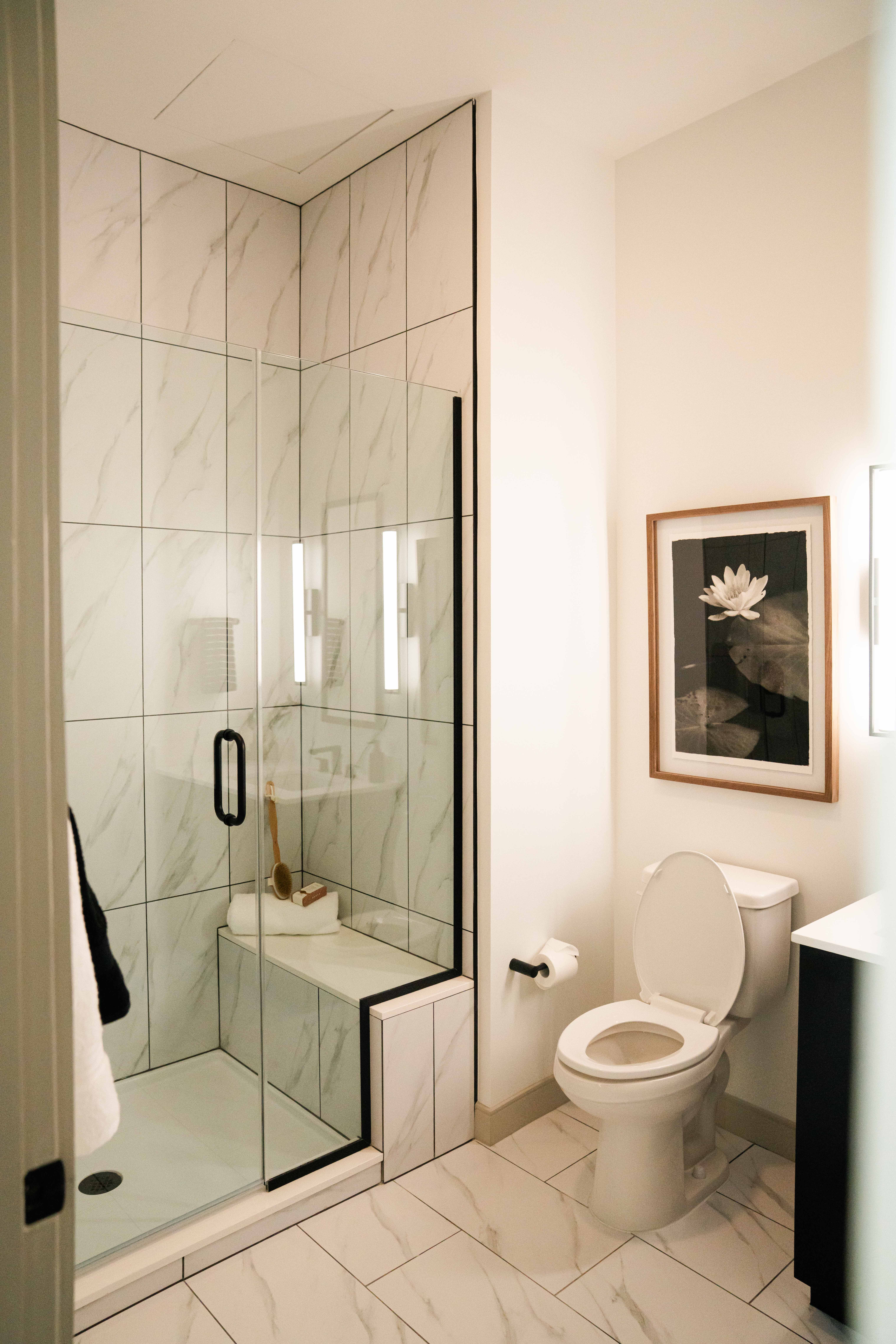 Modern bathroom with glass shower enclosure, black framed door, white marble tiles, toilet with open lid, wall art of a white flower on dark background, and a towel with brush inside shower.