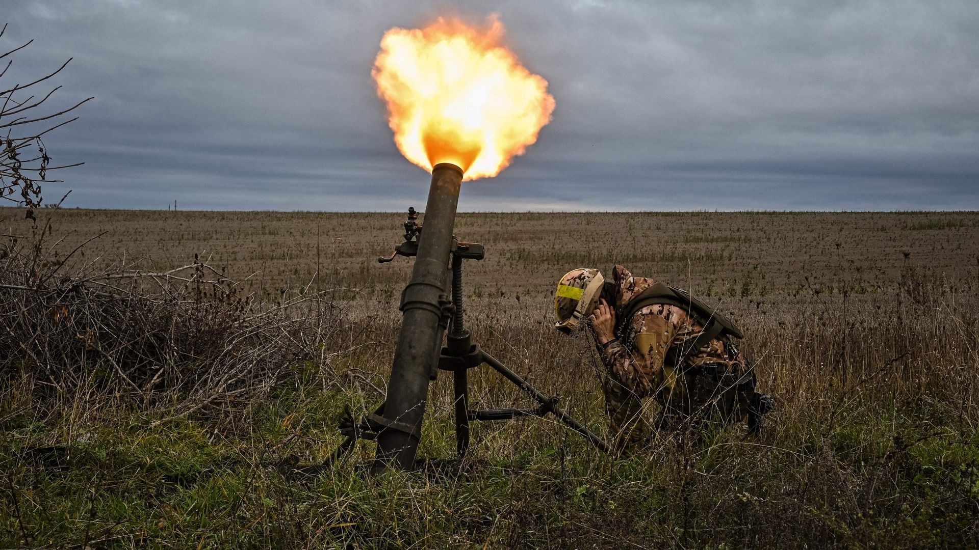 A person kneels and covers their ears as a mortar is fired nearby. The field they are in is brown and green. The sky overhead is blue and grey.