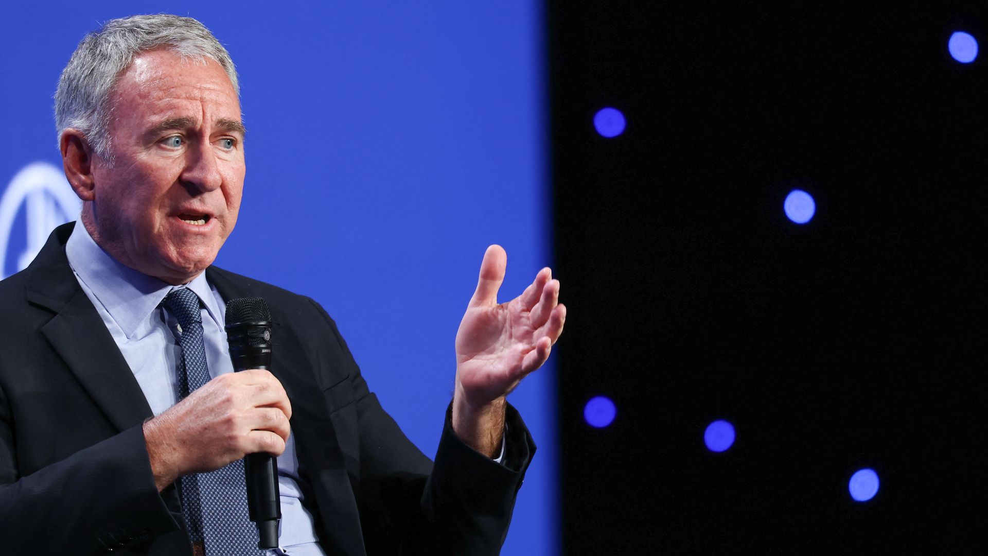 A gray-haired person in a dark suit and light blue shirt speaks into a handheld microphone on a stage, gesturing with his right hand, against a blue backdrop with scattered blue lights.