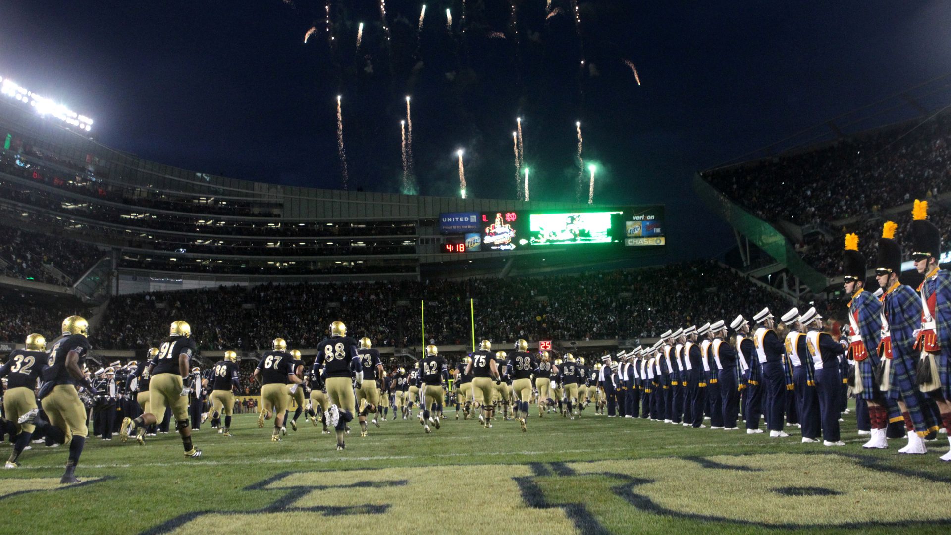 Photo of football players running onto a field as fireworks shoot off in distance.
