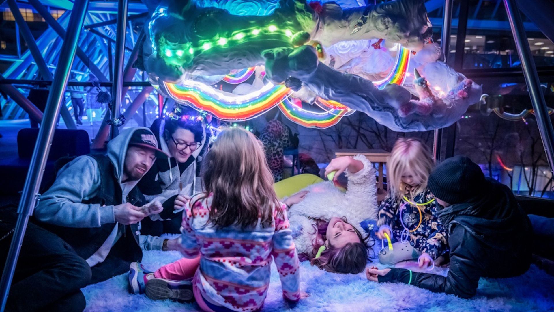 A photo of a family of six, laying underneath a lit-up rainbow cloud art installation.