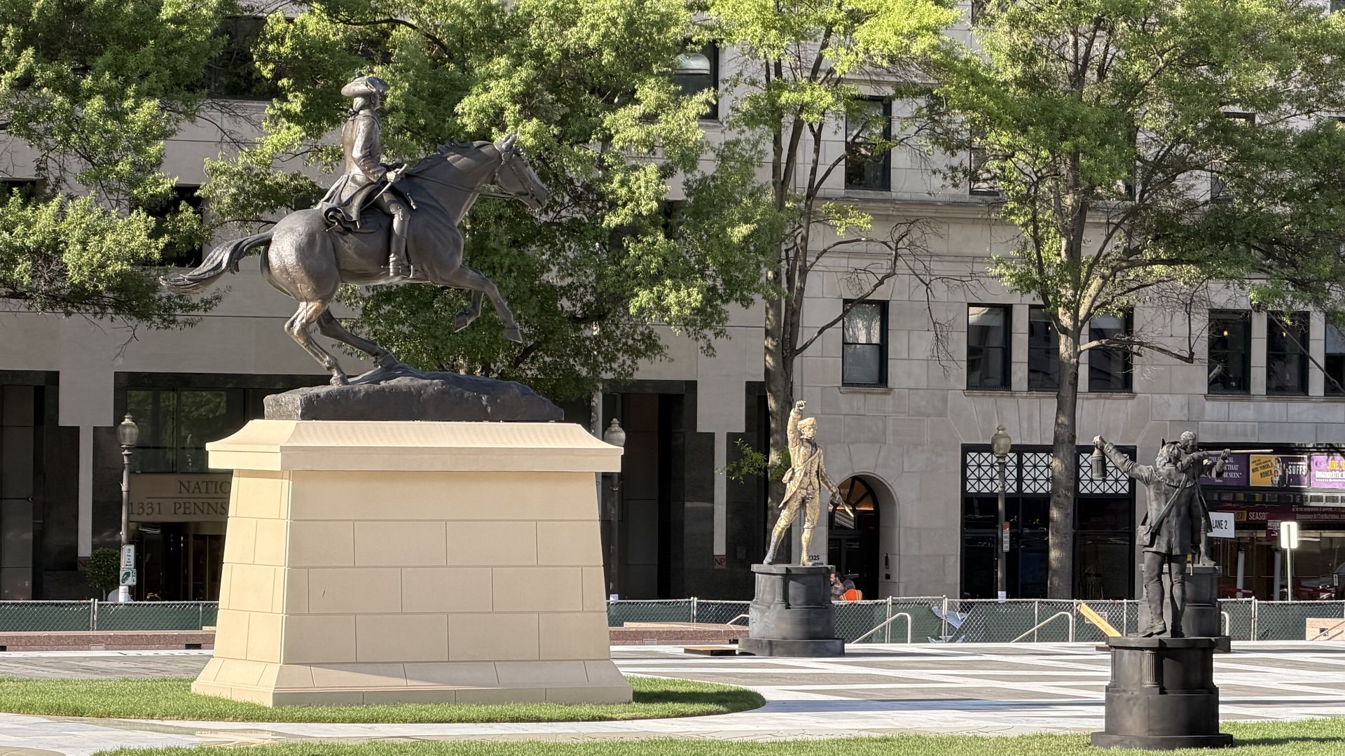 Bronze equestrian statue of a rider on a galloping horse in a plaza, with green trees and a beige office building behind; other statues and a fence in sight.
