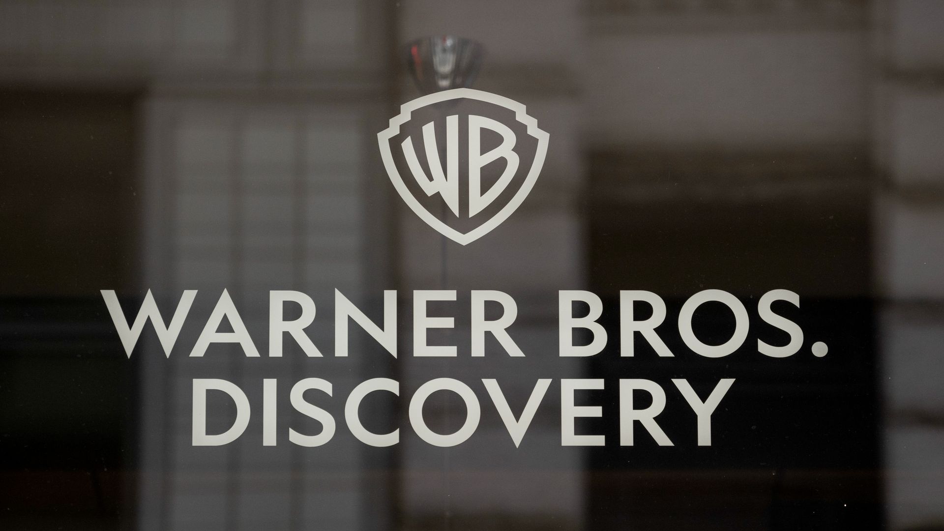 White Warner Bros. Discovery logo and text on a dark, reflective glass surface with a blurred building reflection in the background.