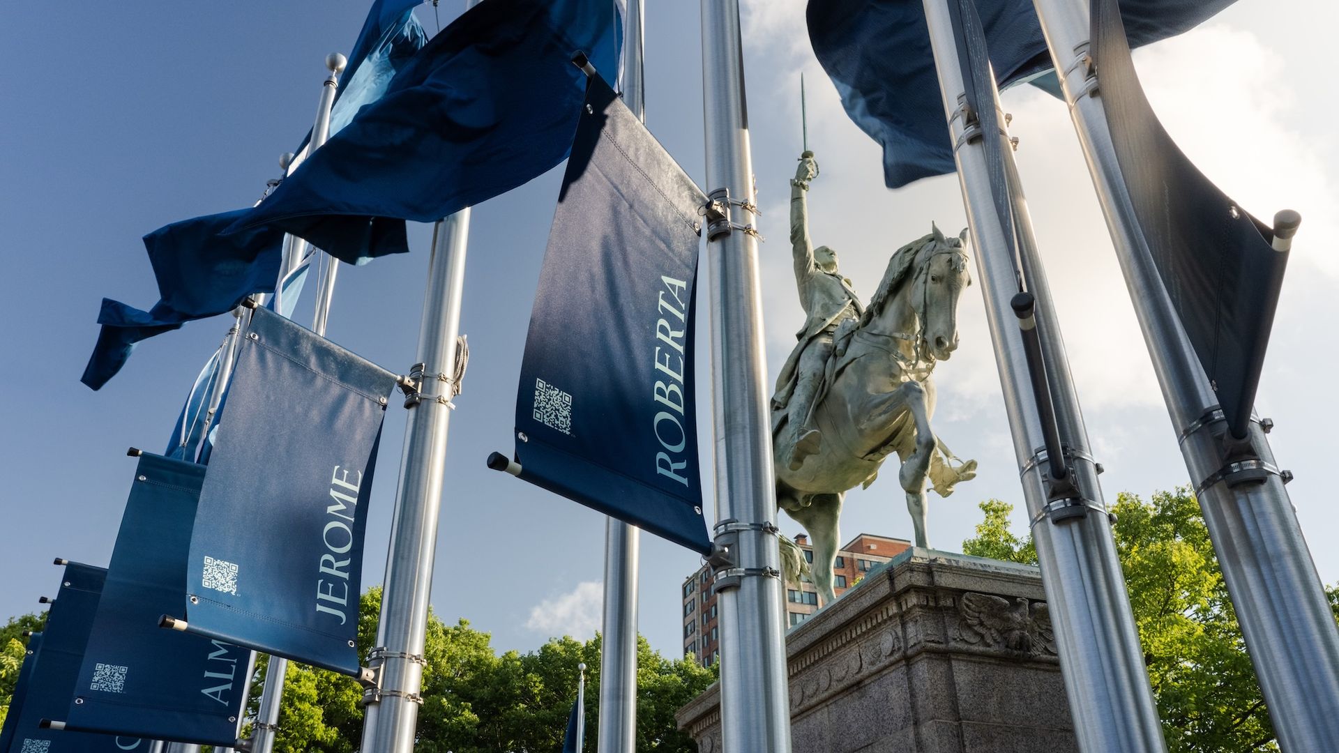 Blue banners hang from tall metal flagpoles around a stone monument with a horse-and-rider statue; trees, a brick building, and a clear blue sky in the background.