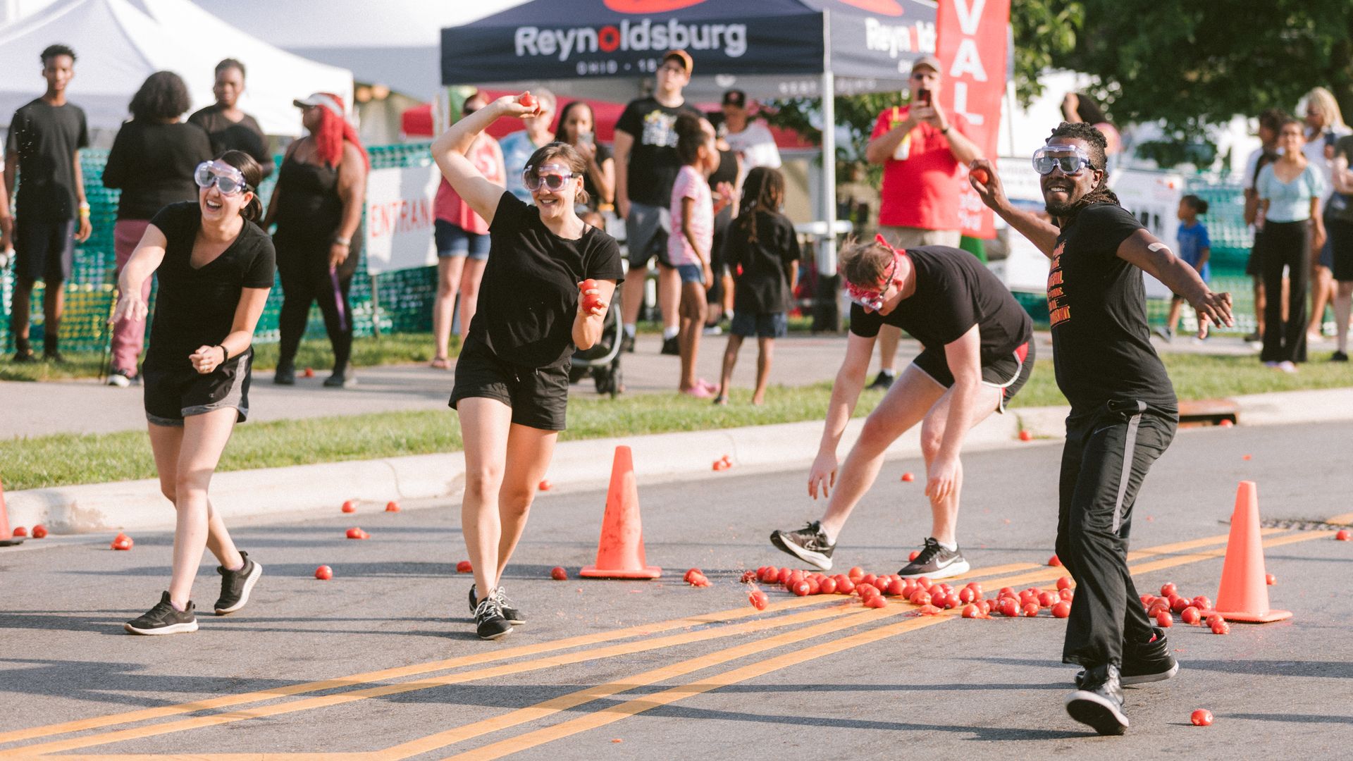 Four people in goggles and black clothing throw tomatoes at each other in a street full of tomatoes and traffic cones