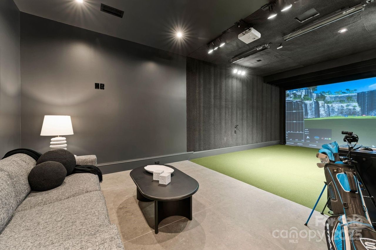 Modern indoor golf simulator room with gray walls, green putting turf, gray sofa with black pillows, a black coffee table, and a golf bag with clubs near a large screen showing a golf course.