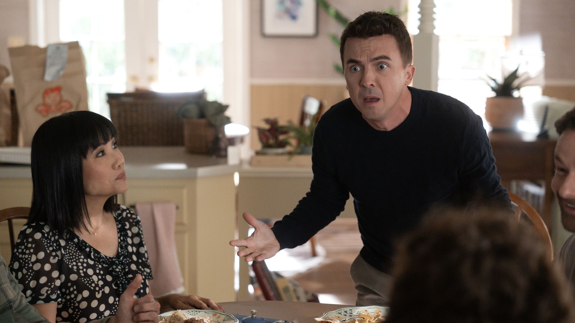 Man in a black shirt leans over a dining table with a shocked expression, gesturing. A woman with black hair in a polka-dot dress looks up as plates of food and blue cups sit on the table.
