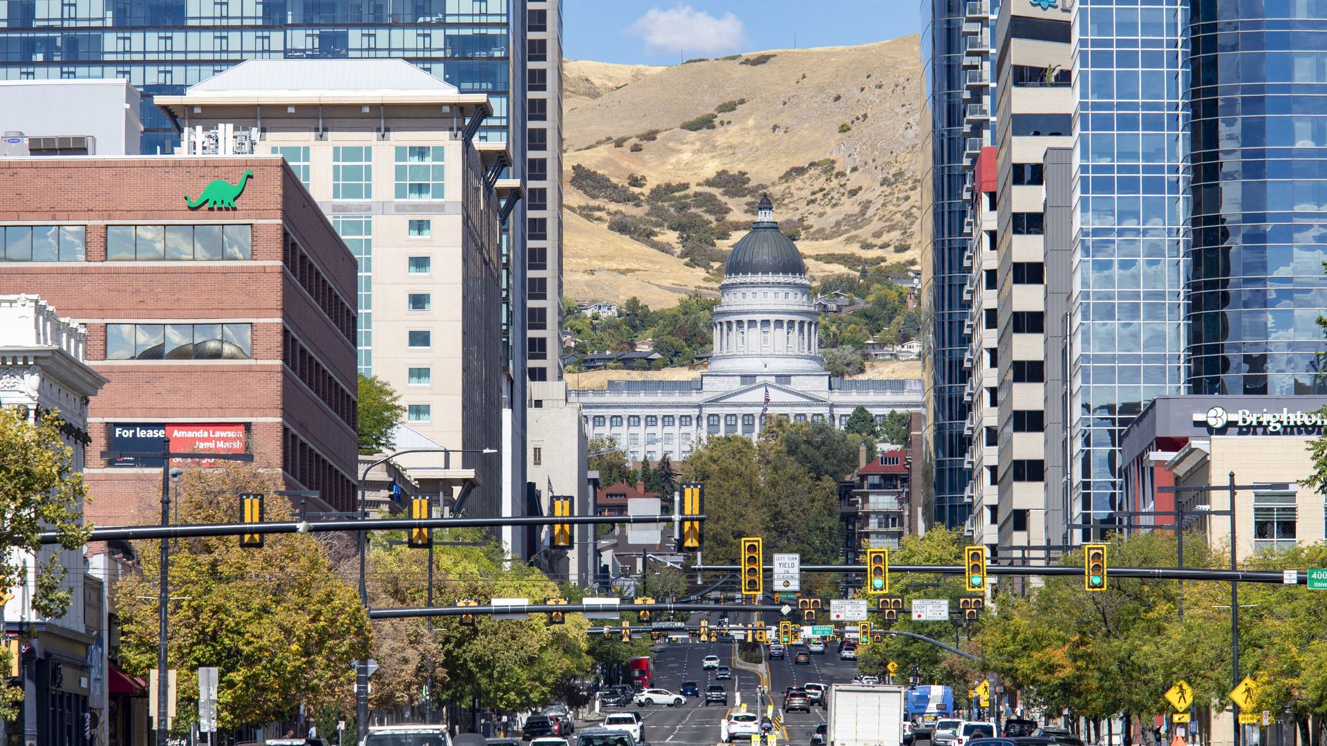 Busy city street flanked by glass office towers; a domed capitol building sits against a hill in the background, with numerous cars and traffic signals in view.