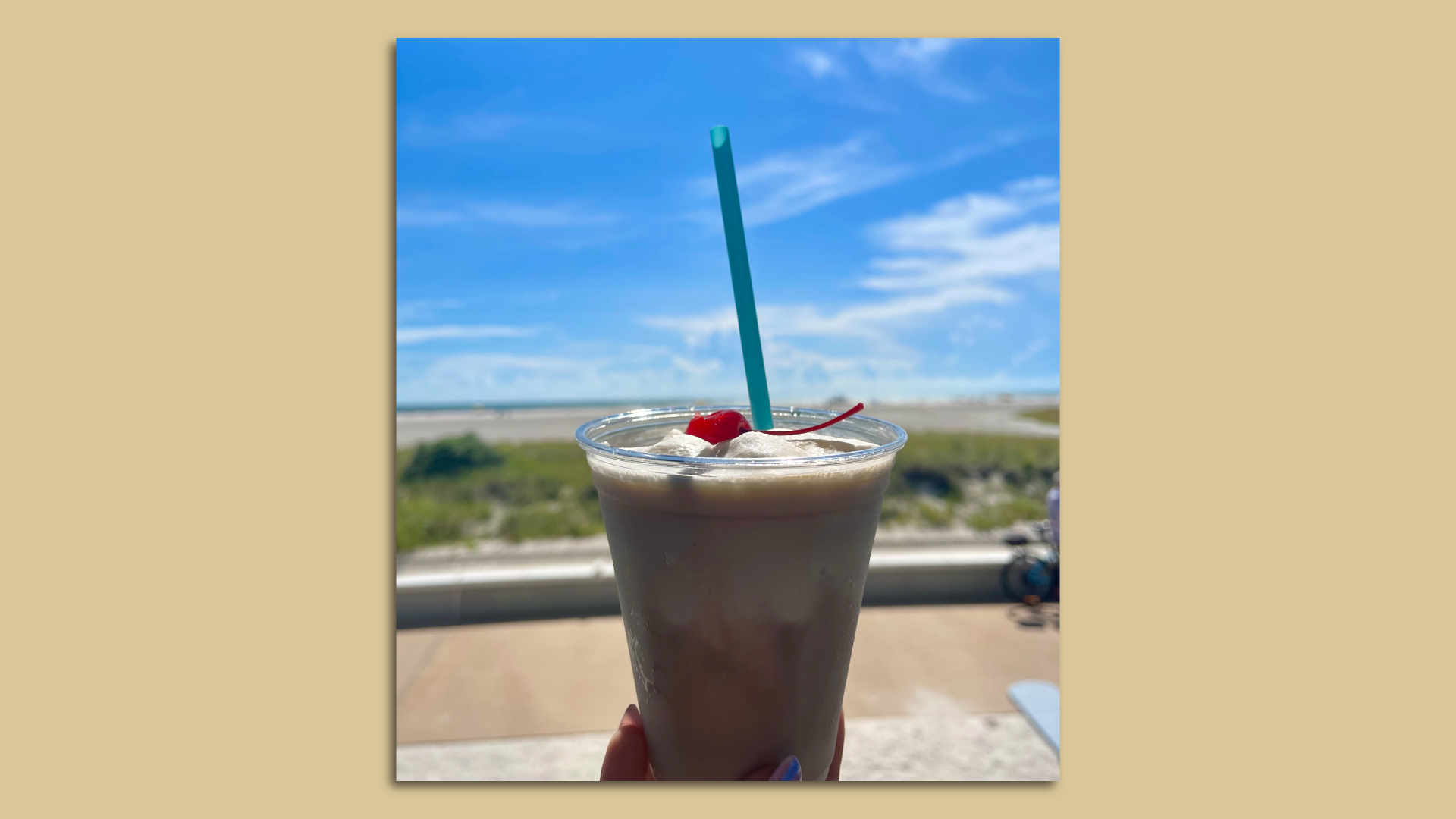 A Bushwacker frozen cocktail in a clear cup with a blue straw, in front of the beach.