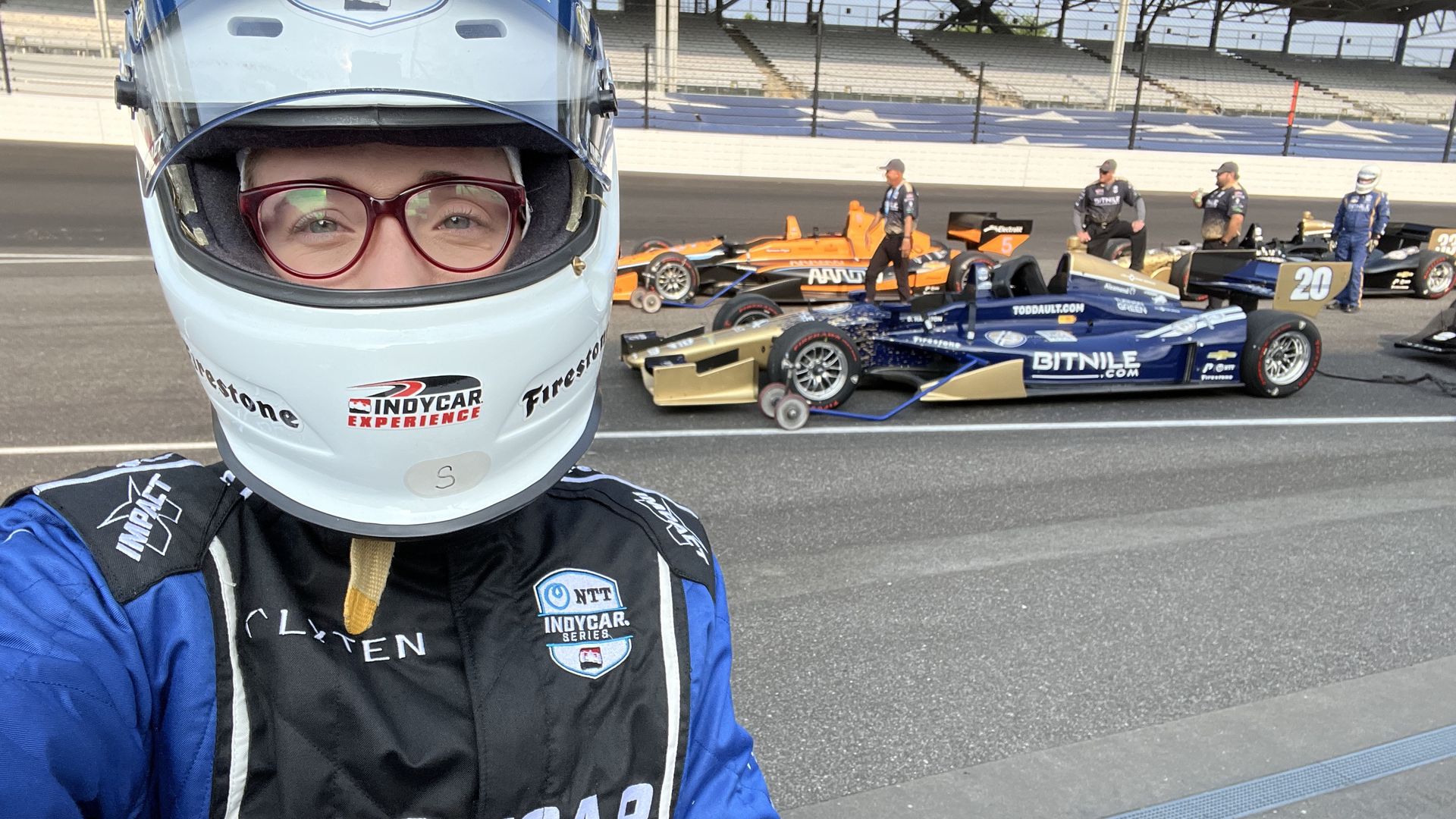 A woman in the foreground wearing a fire suit and helmet, with IndyCars in the background on a race track.
