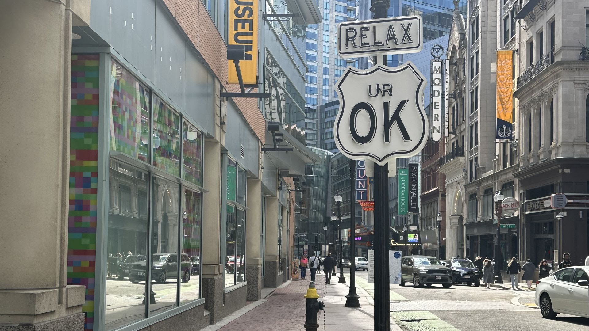 Busy city street with tall buildings and colorful storefronts. A large retro Route 66 shield sign reads "UR OK" and a "RELAX" sign above. Pedestrians walk the brick sidewalk while cars drive by.