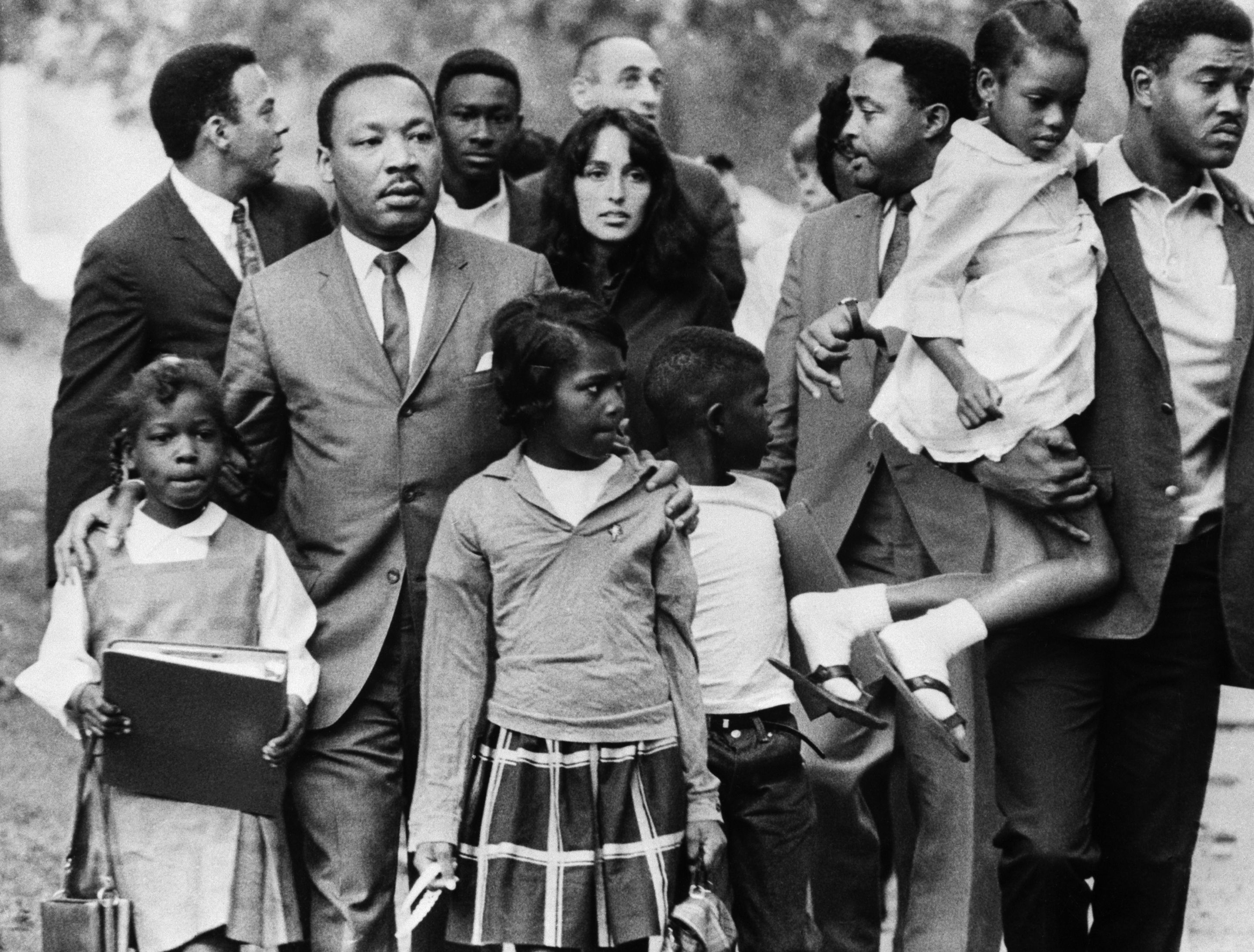 Dr. Martin Luther King, Jr. is shown leading a group of black children to their newly integrated school in Grenada, Mississippi, escorted by folk singer Joan Baez and two aides.