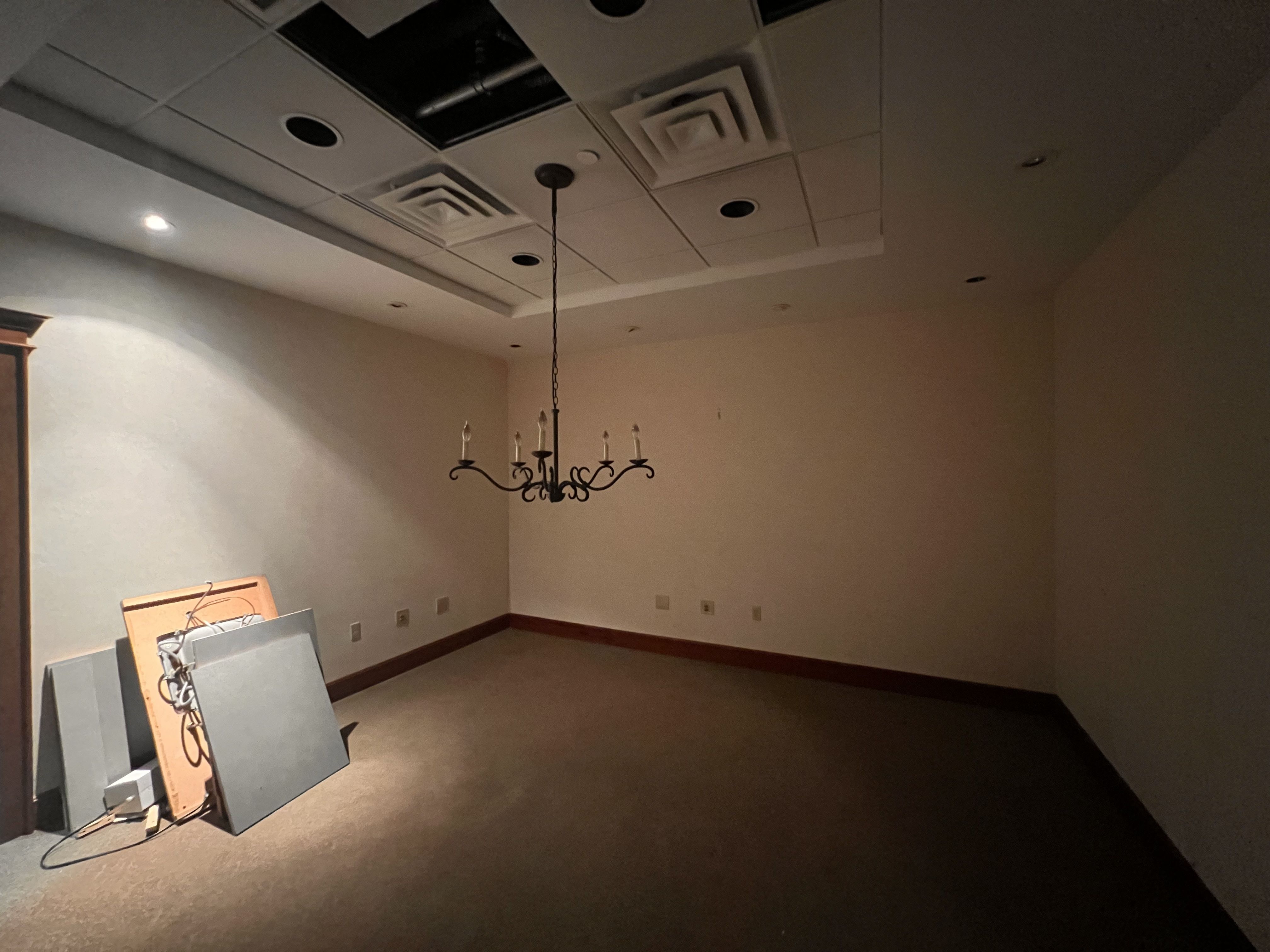 Dimly lit, empty room under renovation. Beige walls, brown baseboard. A black ornate chandelier hangs from a chain from a ceiling with missing panels; leaning boards rest along the left wall.
