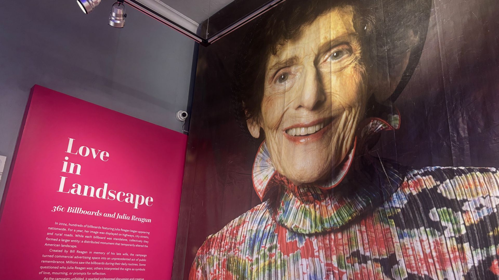 Interior wall mural of a large, smiling elderly woman with a colorful ruffled collar. A pink panel on the left reads "Love in Landscape" with a spotlight overhead.