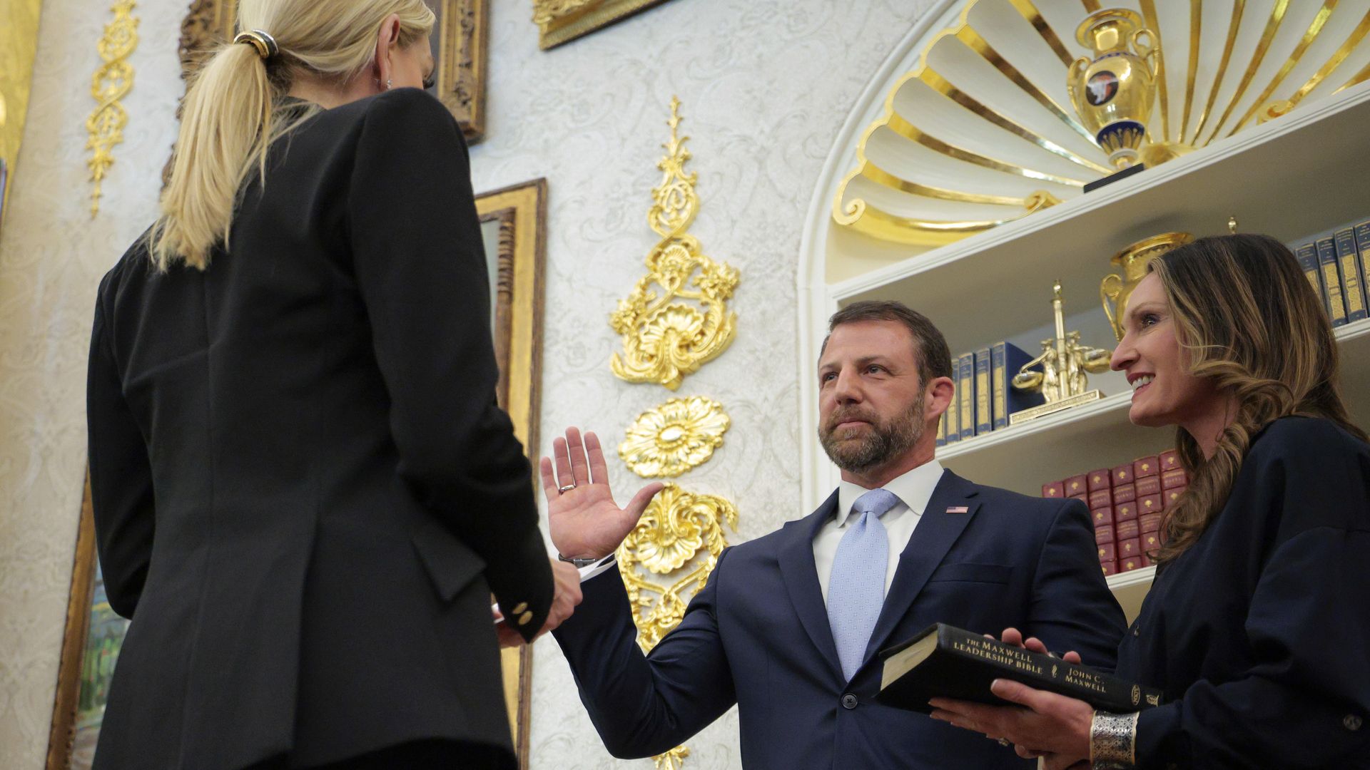 Markwayne Mullin — wearing a dark blue suit, a light blue tie and a white collared shirt — holds up his hand as he's sworn in by Attorney General Pam Bondi, who is facing him wearing a black jacket. His wife, wearing black and holding a Bible, stands next to him.
