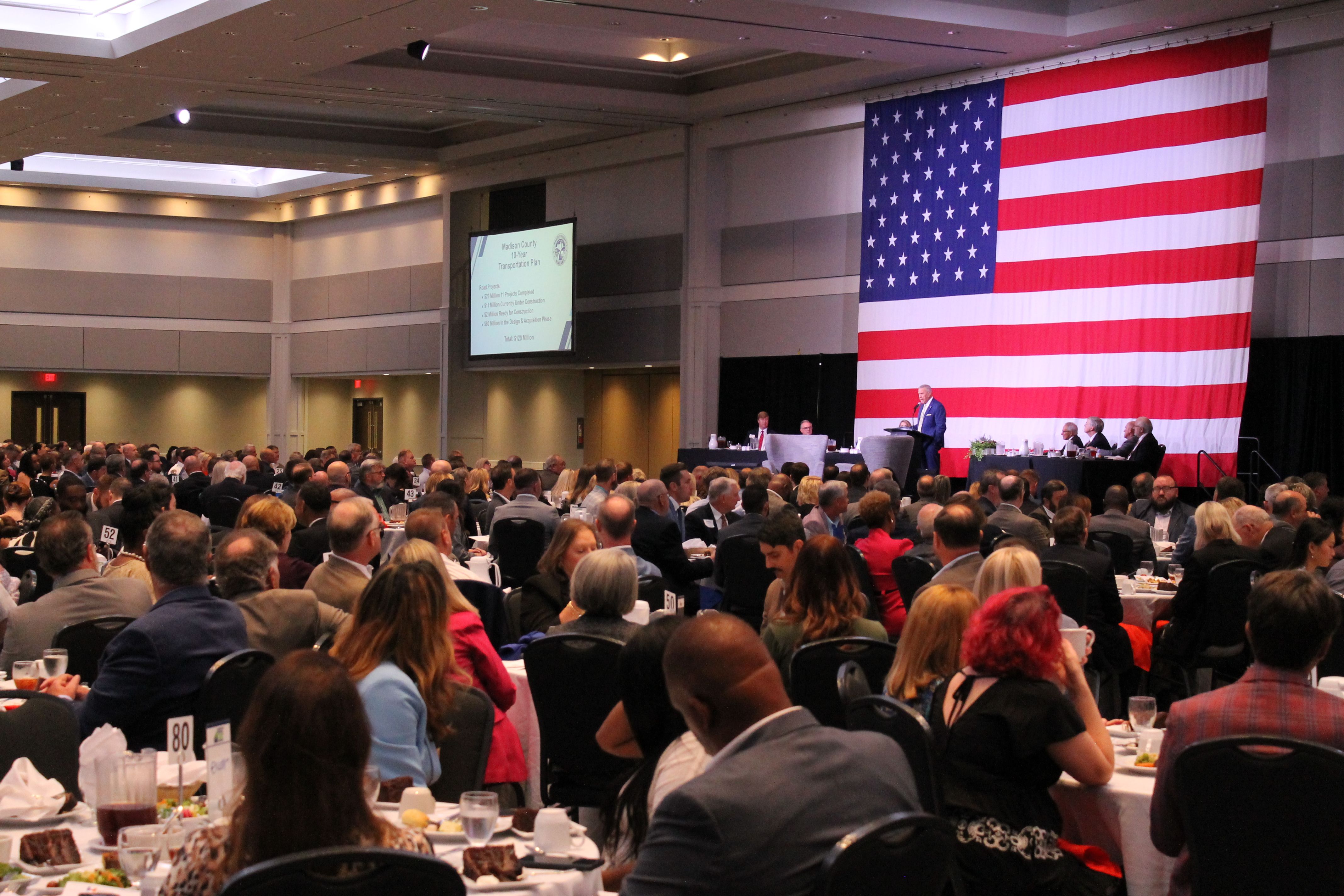 Large audience seated at round tables in a conference hall, listening to a speaker on stage with a large American flag backdrop and a presentation screen about Madison County's 10-year Transportation Plan.