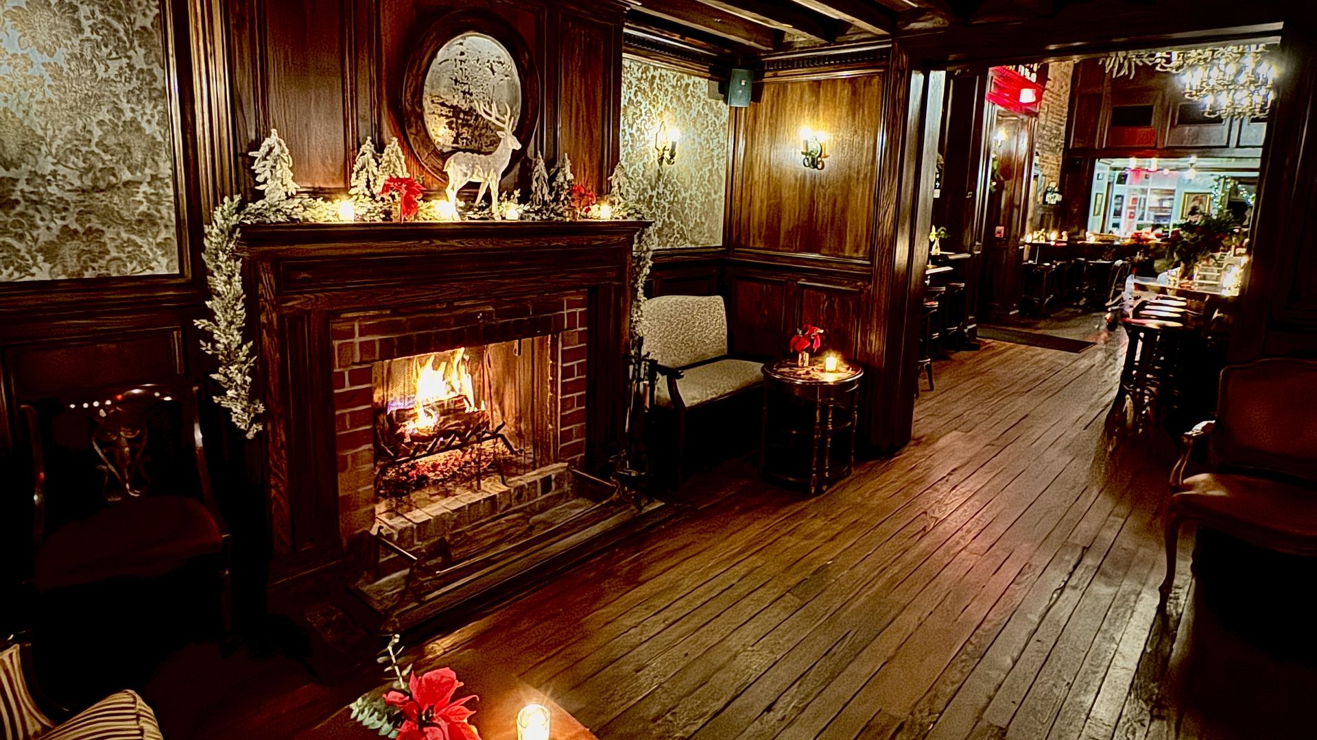 Cozy room with wooden floors and paneling, lit fireplace decorated with white trees and reindeer figure, candles and red poinsettias on tables, and warm wall sconces.