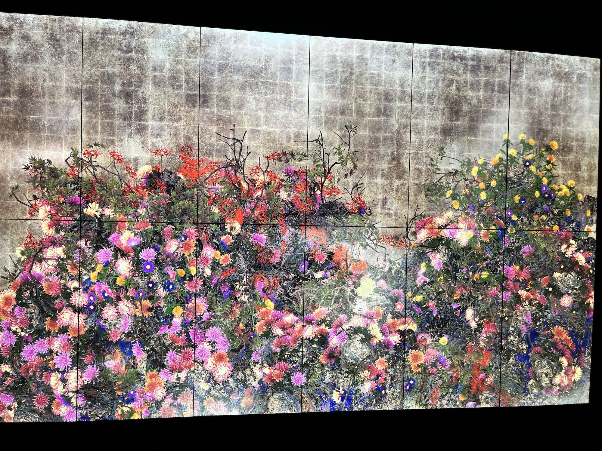 Wide mural shows a dense bouquet of multi-colored flowers along the bottom, including pink, red, yellow and purple, against a weathered gray tiled wall with panel seams and a dark ceiling above.
