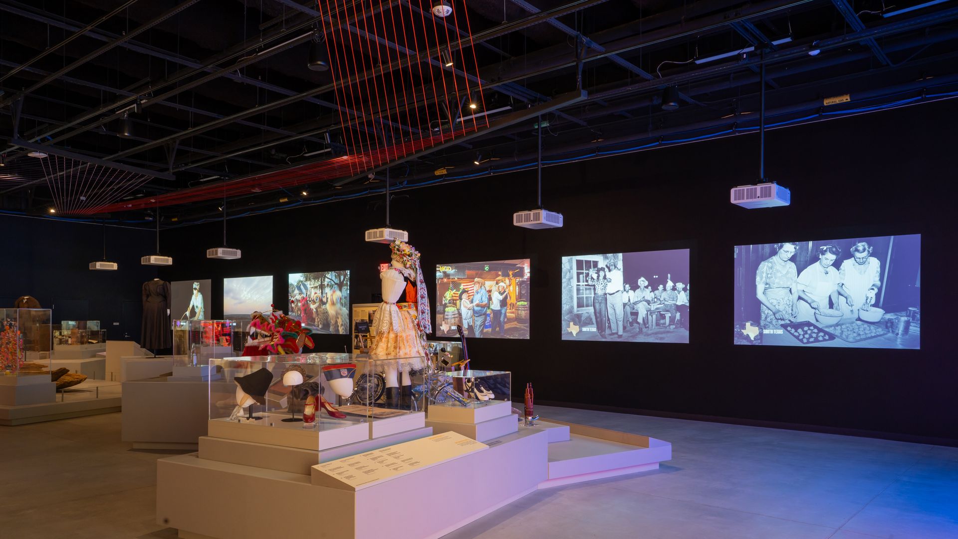 Museum exhibit with traditional and cultural artifacts in glass cases, a mannequin in a colorful dress, and five projected images on a black wall, including black-and-white photos of people.
