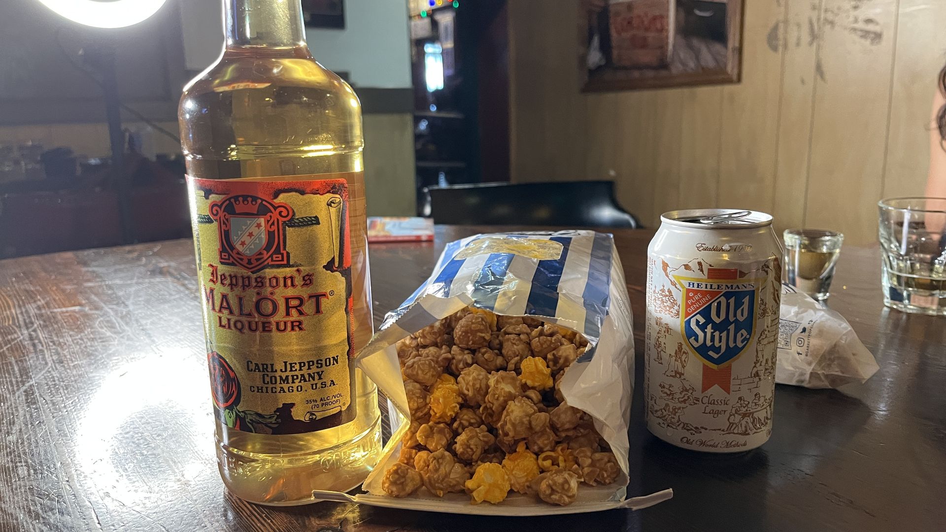 Close-up of a bottle of Jeppson's Malört liqueur on a wooden table beside a can of Old Style beer, a foil-wrapped snack bag, and popped corn; ring light and bar in background.