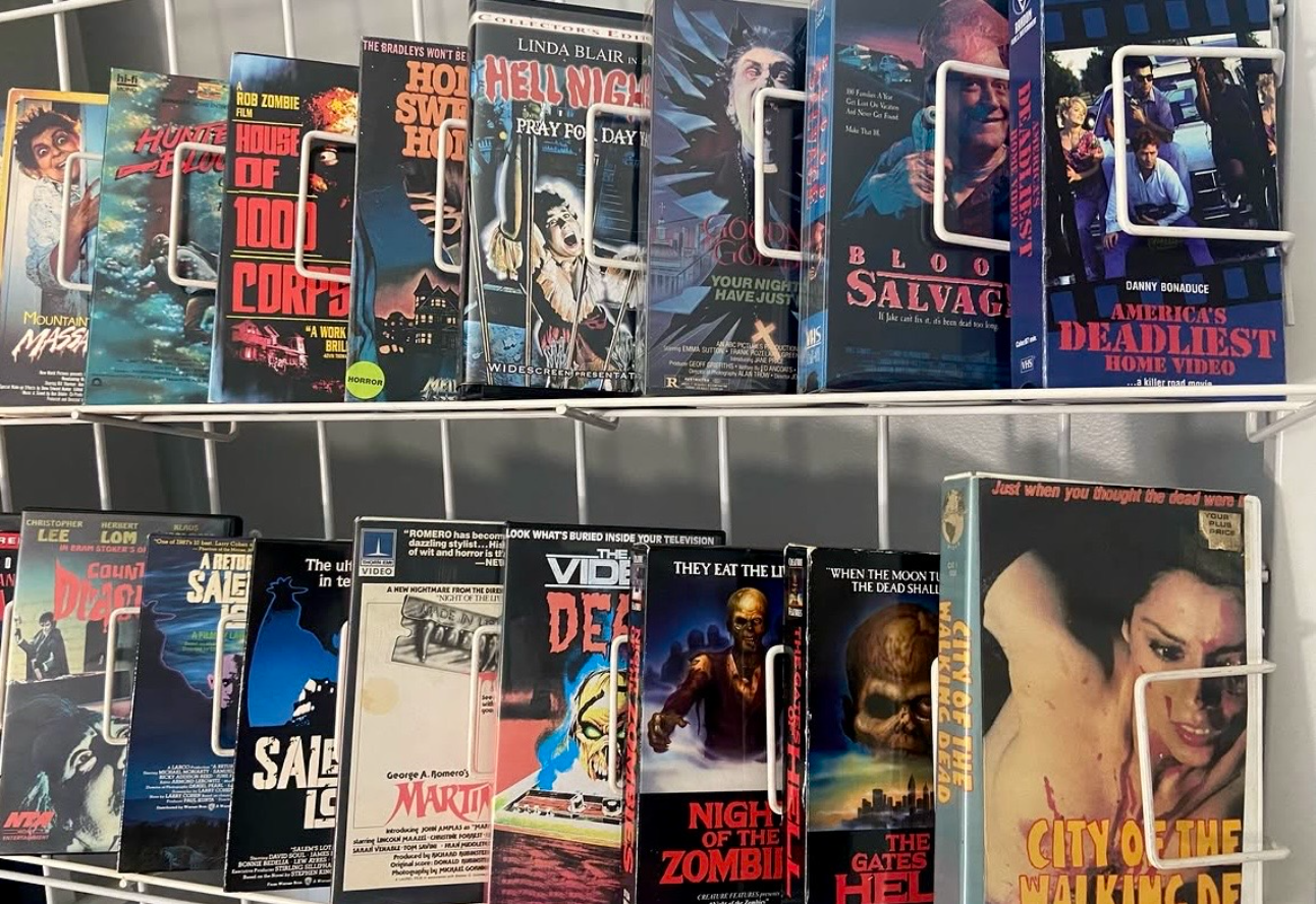 Two-tier metal rack filled with colorful vintage horror VHS covers, bold red, blue, and black artwork. Notable titles include "House of 1000 Corpses" and "America's Deadliest Home Video".