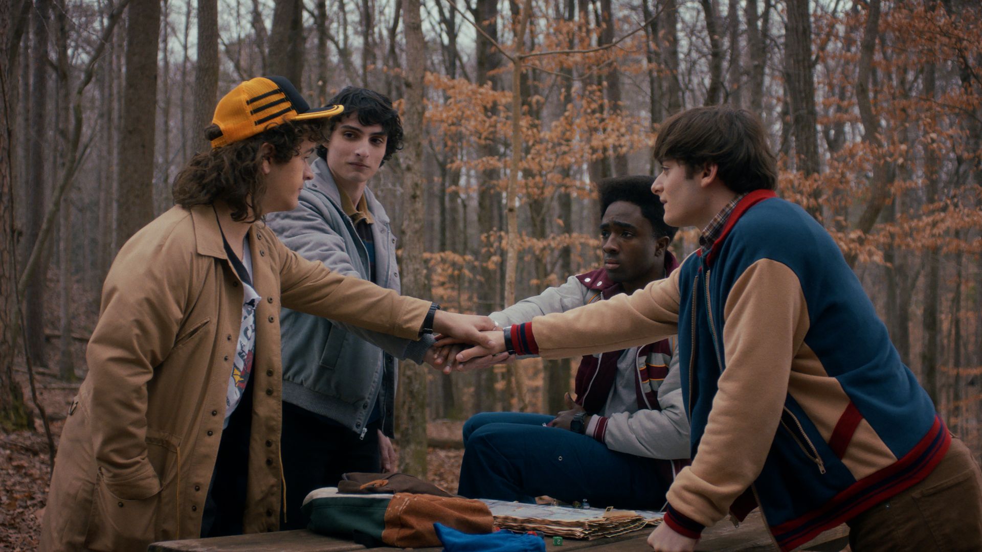 Four teenage boys stand and sit around a wooden picnic table in a forest with bare trees, all placing their hands together in a group gesture, wearing jackets and casual clothes.