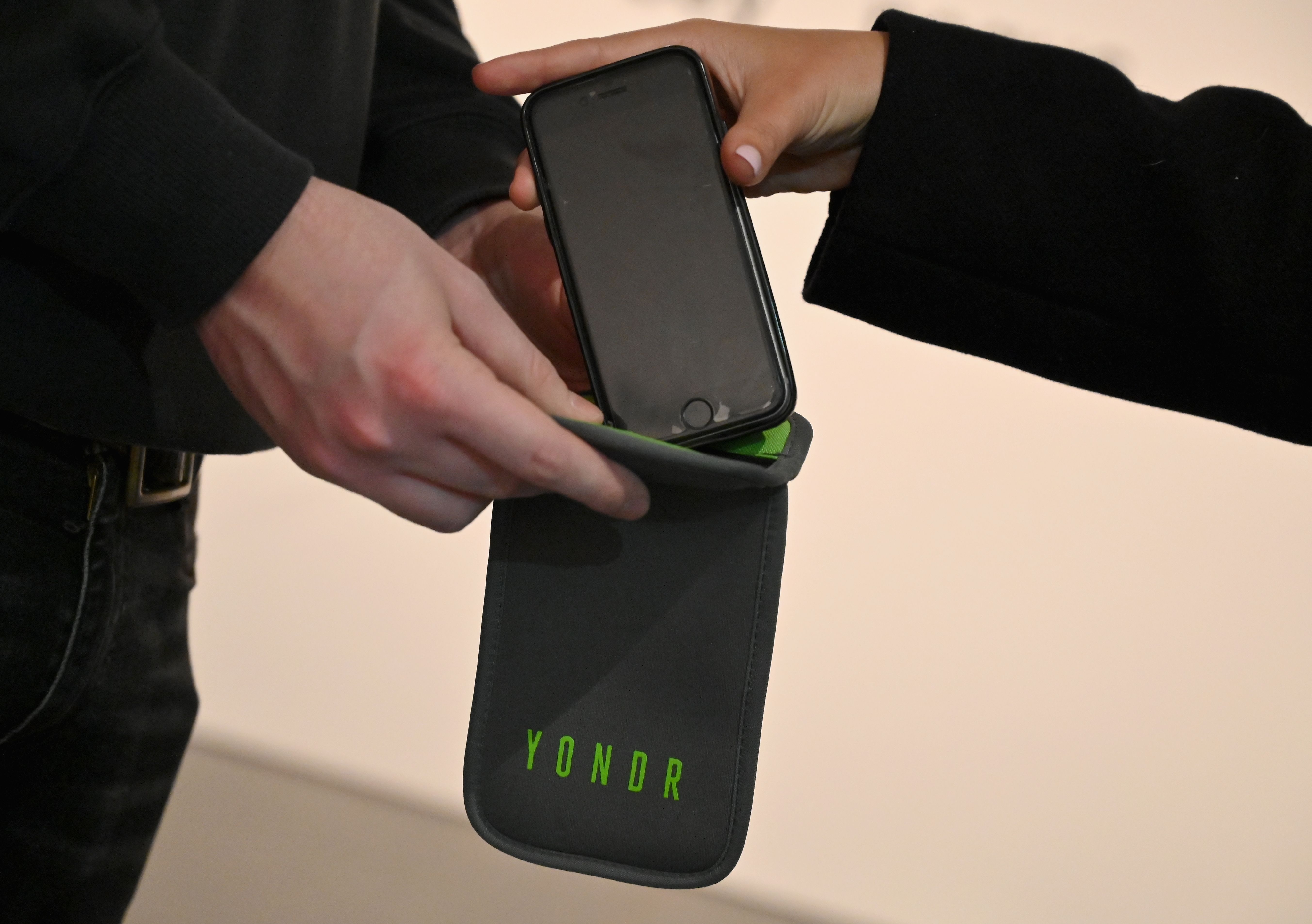 Two people exchanging a smartphone being placed inside a black pouch with green interior labeled "YONDR" in green letters.