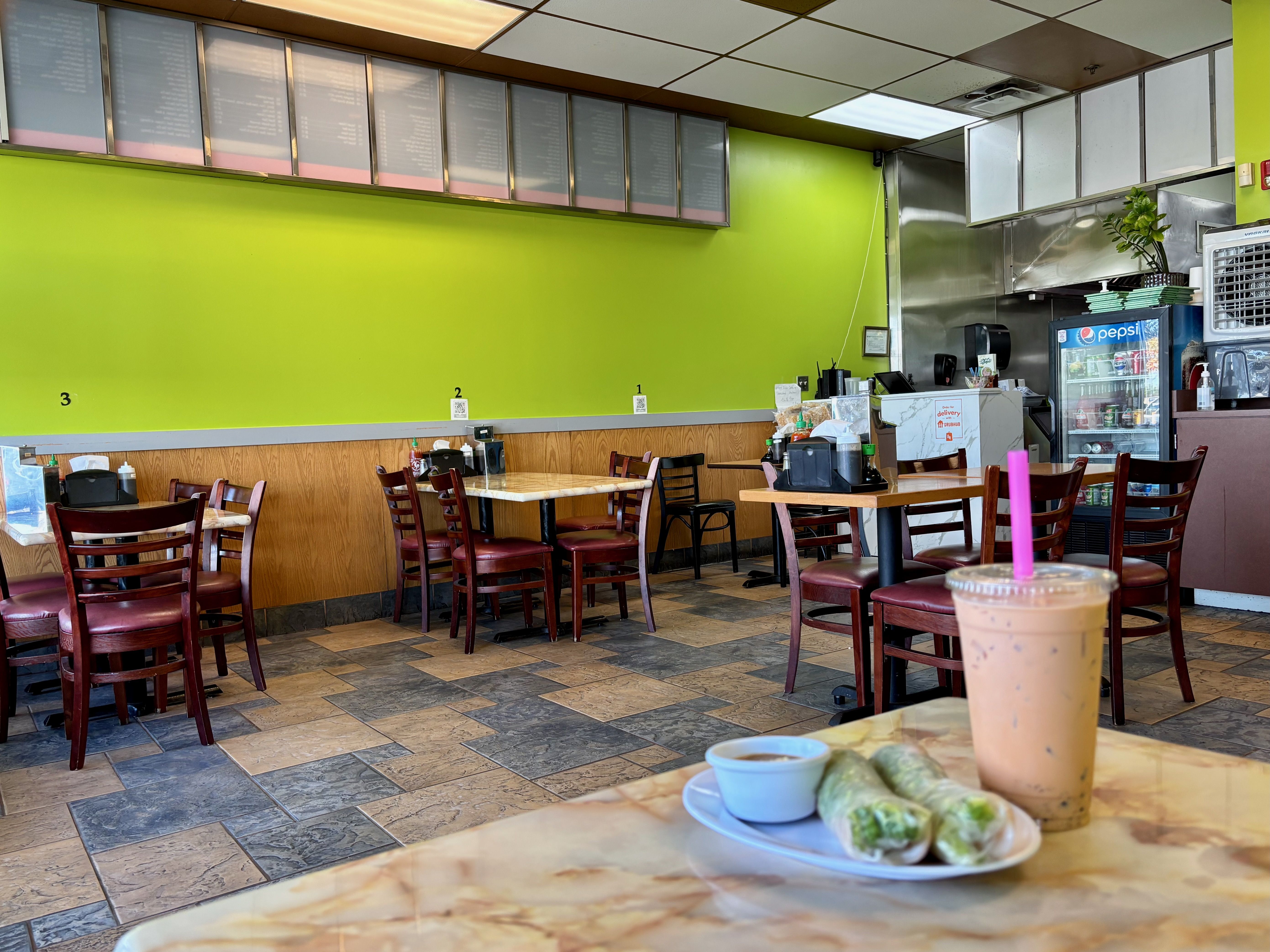 Casual eatery with lime green wall, wooden chairs, and tiled floor. Plate with spring rolls and dipping sauce, and iced drink with pink straw in foreground.