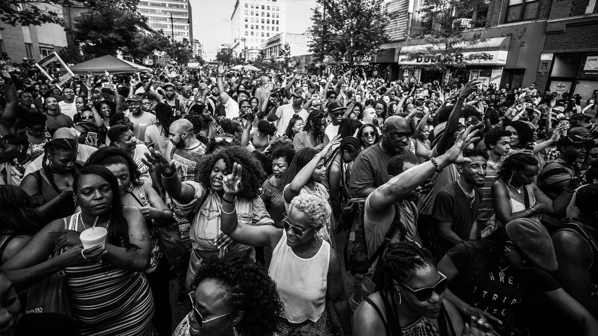 Black-and-white image of a large, diverse crowd with raised hands filling a city street, suggesting a protest or celebration, with buildings lining the block.