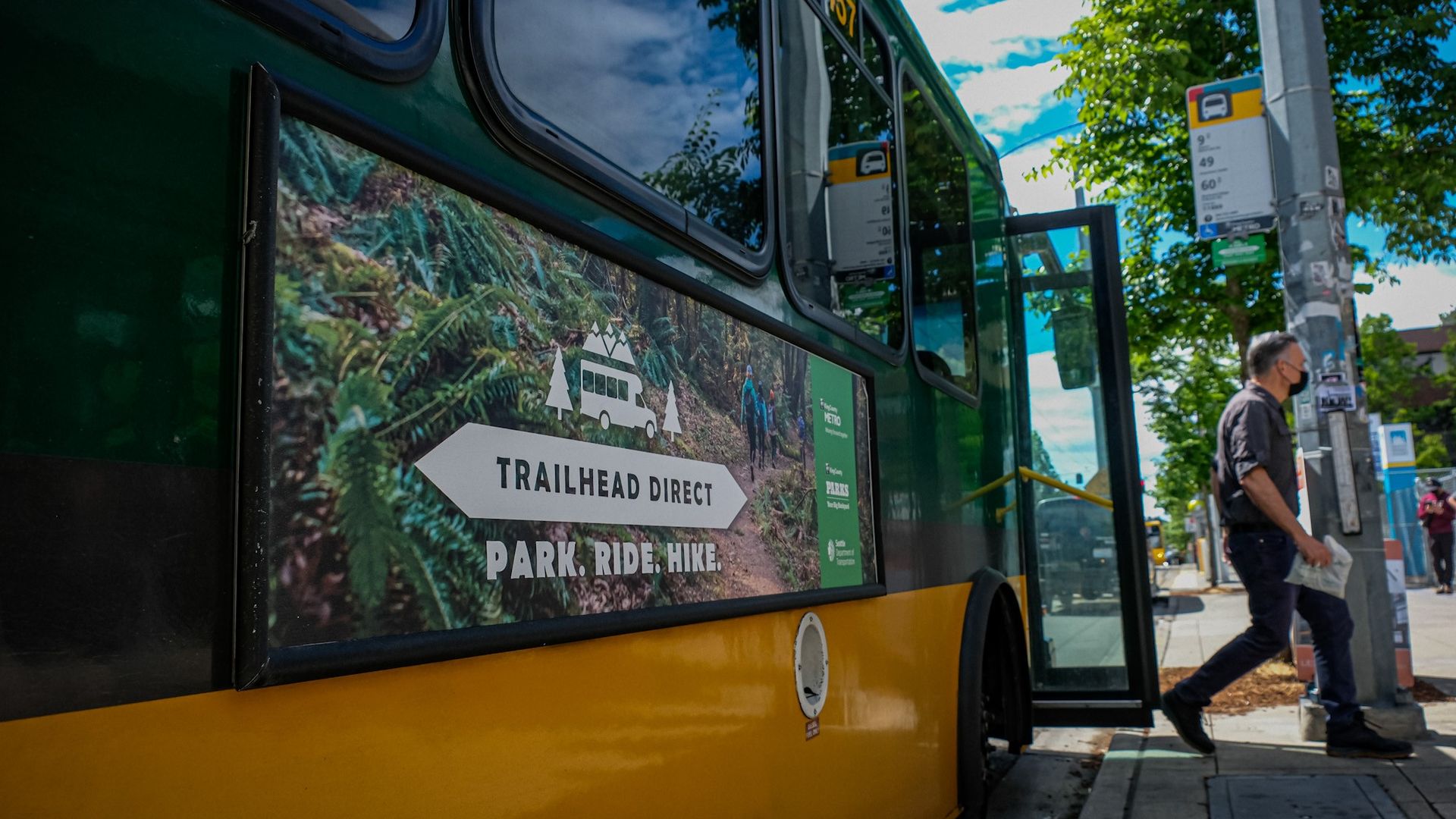 A green and yellow King County metro bus with a sign that says "Trailhead Direct," and a man getting off of the bus onto a curb.