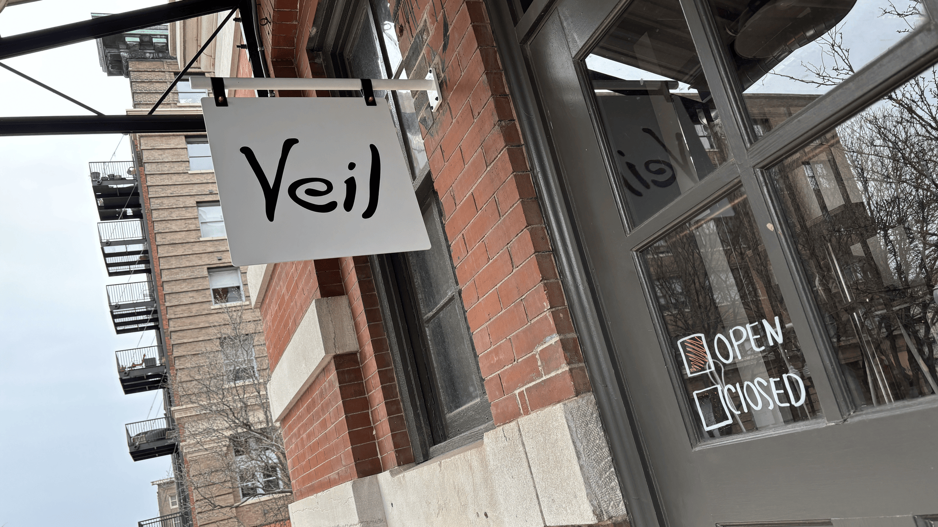 White sign with black text "Veil" hanging outside a brick building. A door with glass panels shows a marker-written sign indicating the store is open.