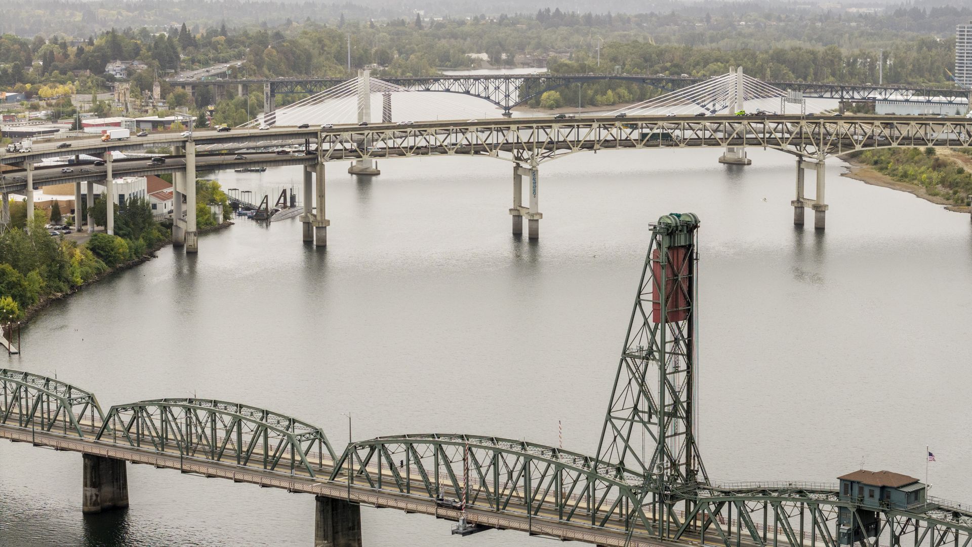 Aerial view of multiple bridges spanning a river, including a green steel truss drawbridge with a tall lift tower in the foreground and cable-stayed and cantilever bridges in the background amid trees and hills.