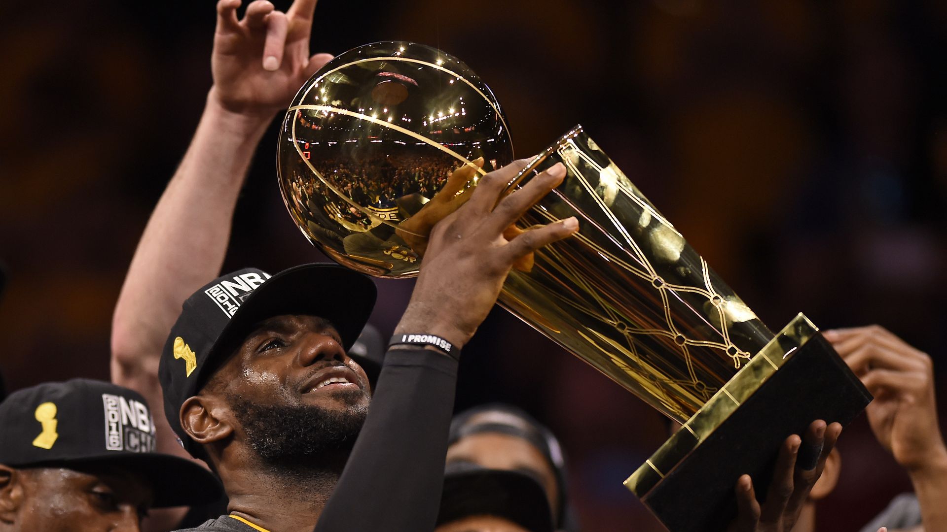 A man wearing a dark cap lifts a large golden glass NBA Championship trophy overhead, its reflective surface showing a cheering crowd and bright stadium lights as celebration surrounds him.