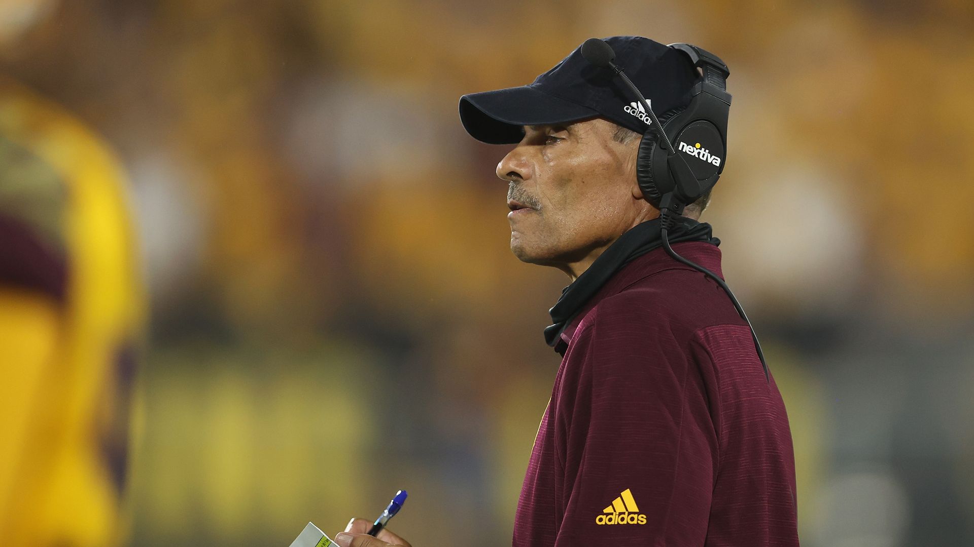 A man holding a notepad and wearing a headset and maroon polo on the sideline of a football field. 